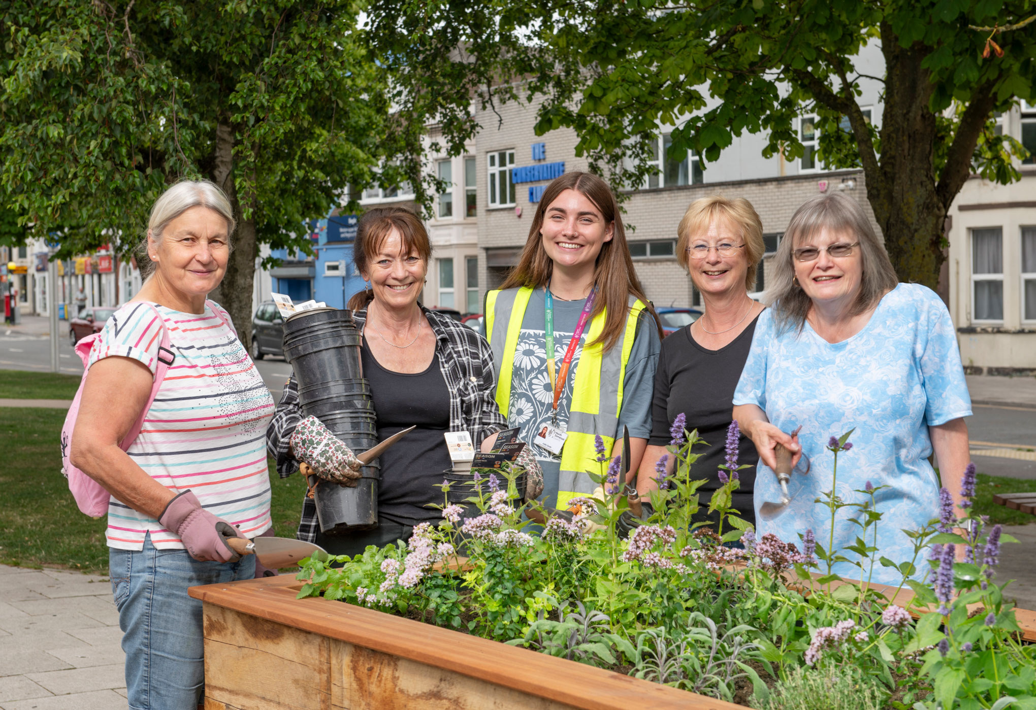 A group of women with gardening tools pose by a large wooden planter on a town street.