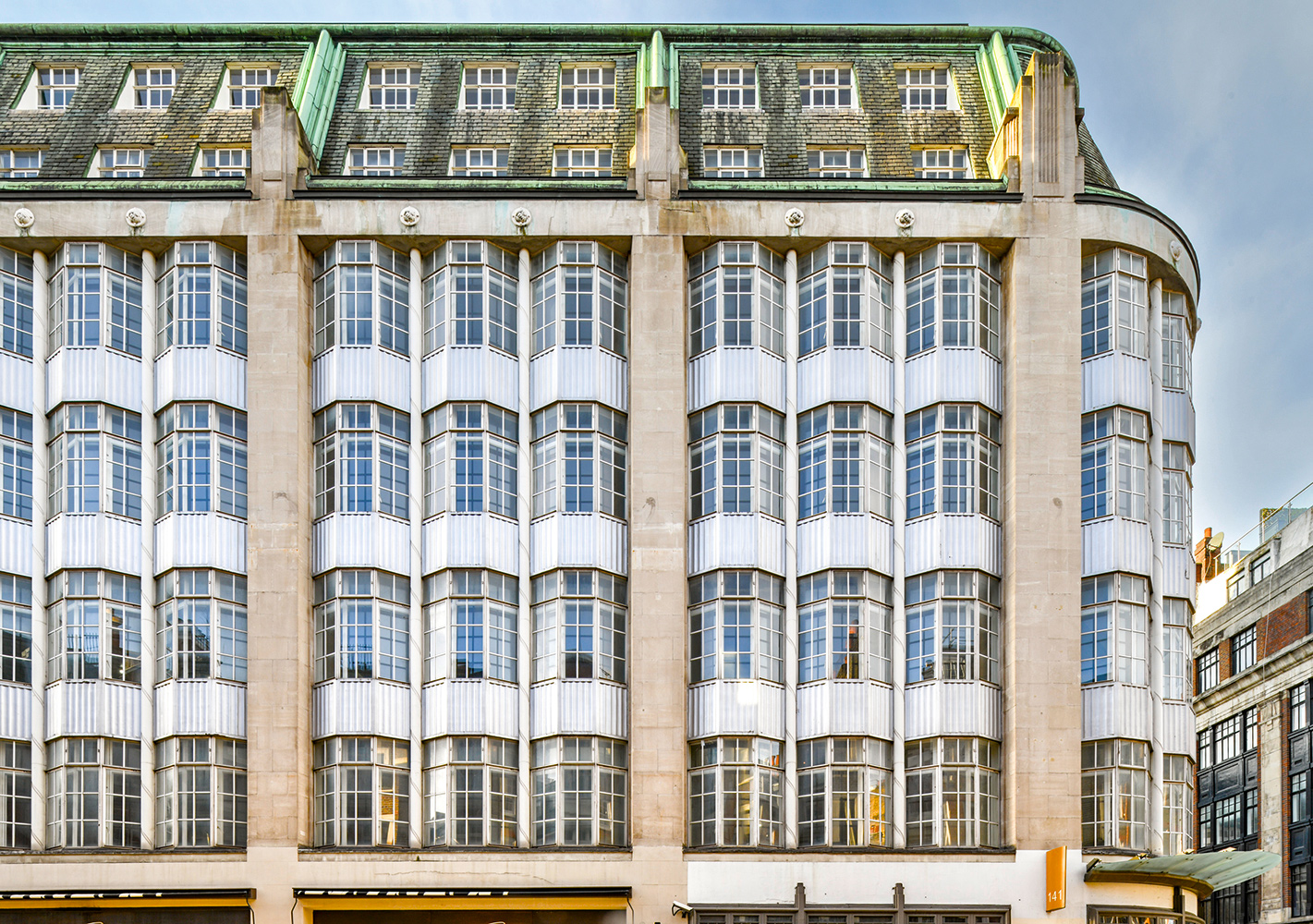 Architectural photo of a large urban Art Deco building frontage. The building is at least 7 storeys high, with a green-glazed brick roof. 