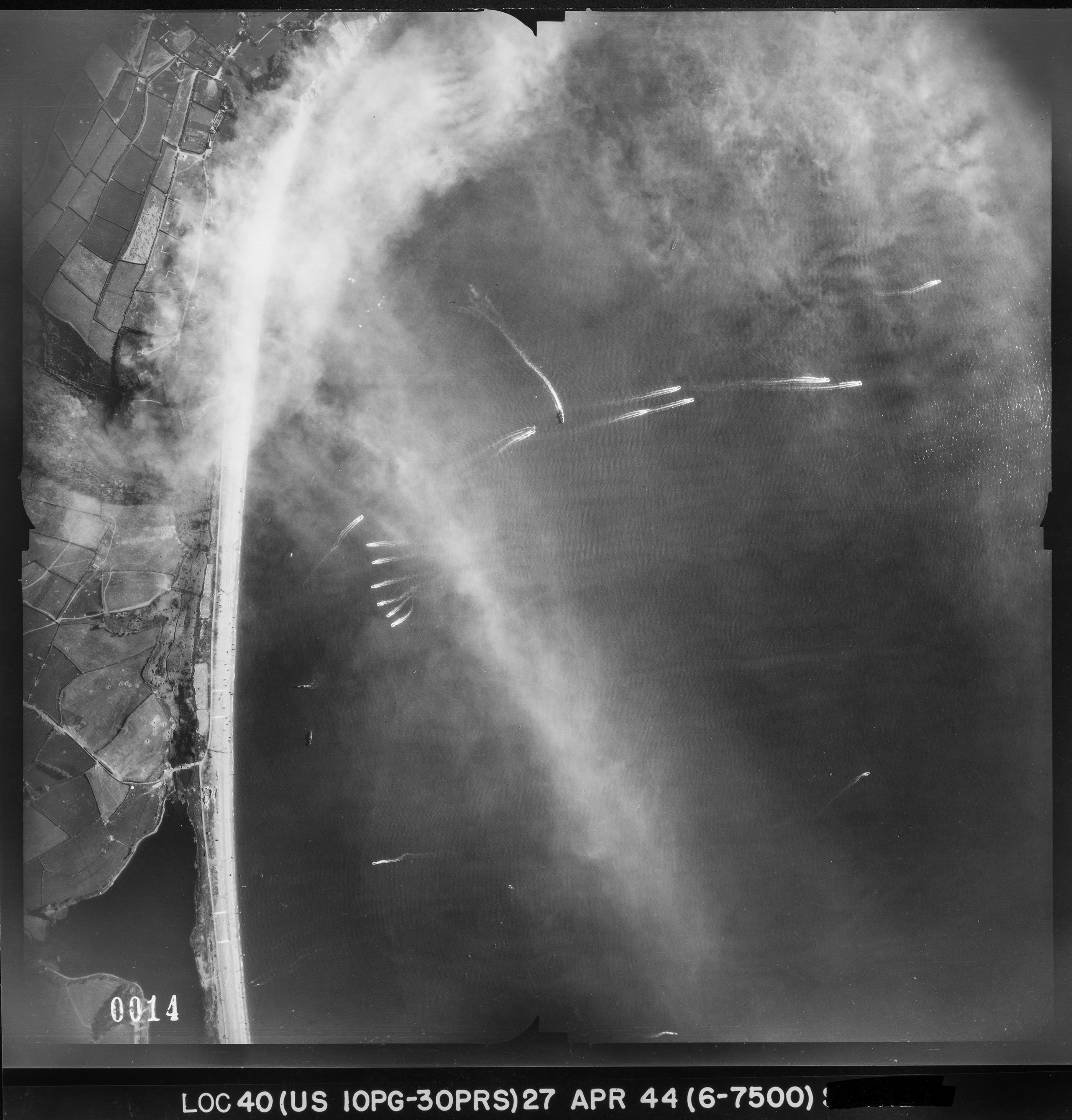 Detail from a black and white vertical aerial photograph, showing ships and their wake on water adjacent to a strip of land. The land is to the left edge of the image and includes a narrow strip of beach next to a patchwork landscape of fields and roads.