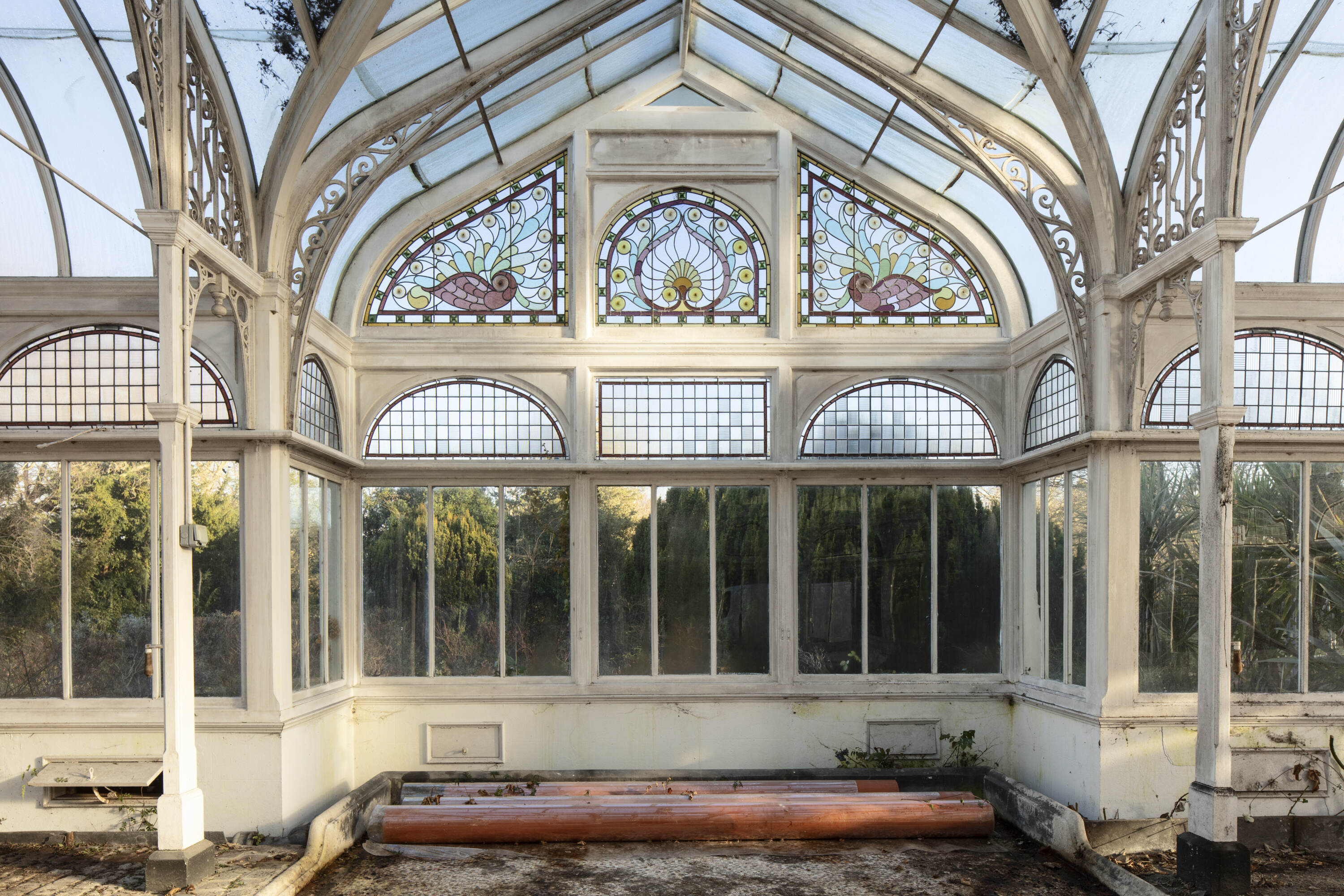 A white framed conservatory with decorative scrolled ironwork and, above plain glass windows, three beautifully decorated panels showing birds and a central peacock feather design.