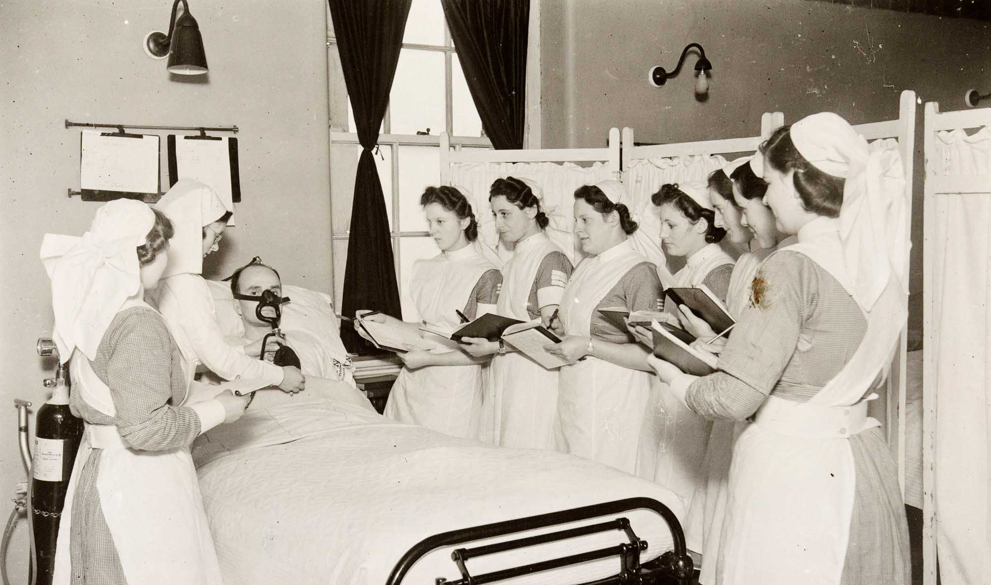 A black and white photograph of student nurses taking notes around a patient's bed, whilst a sister tutor explains the use of an oxygen mask