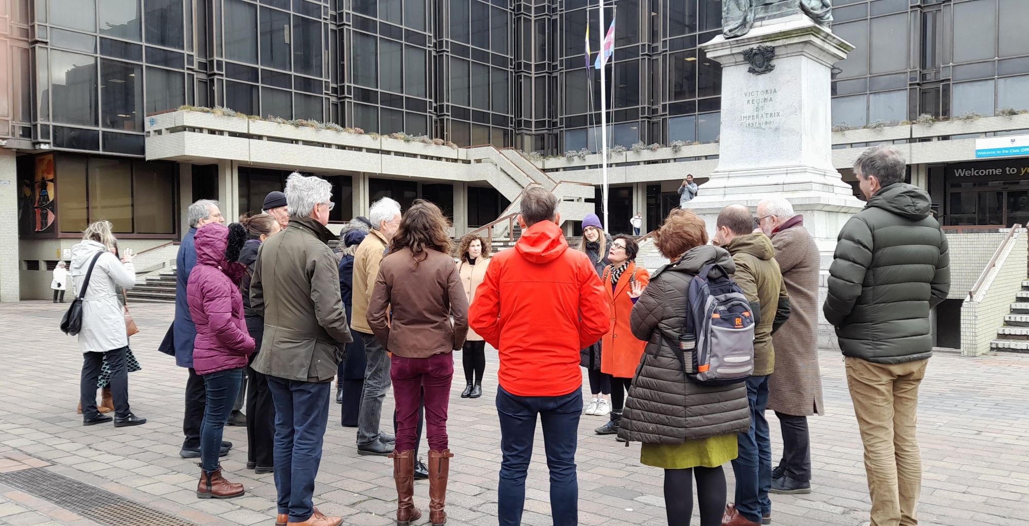 A group of people in warm coats stand listening to a woman speaking.