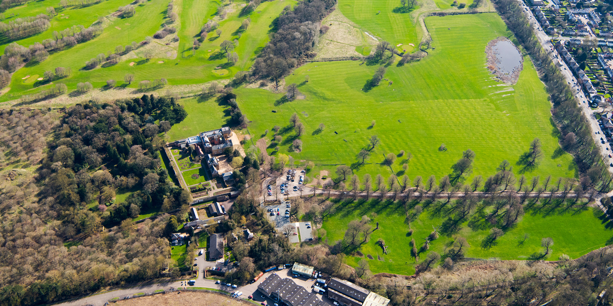 A flooded green turf field surrounded by trees, a golf course, a tree-lined drive, and a busy main road.