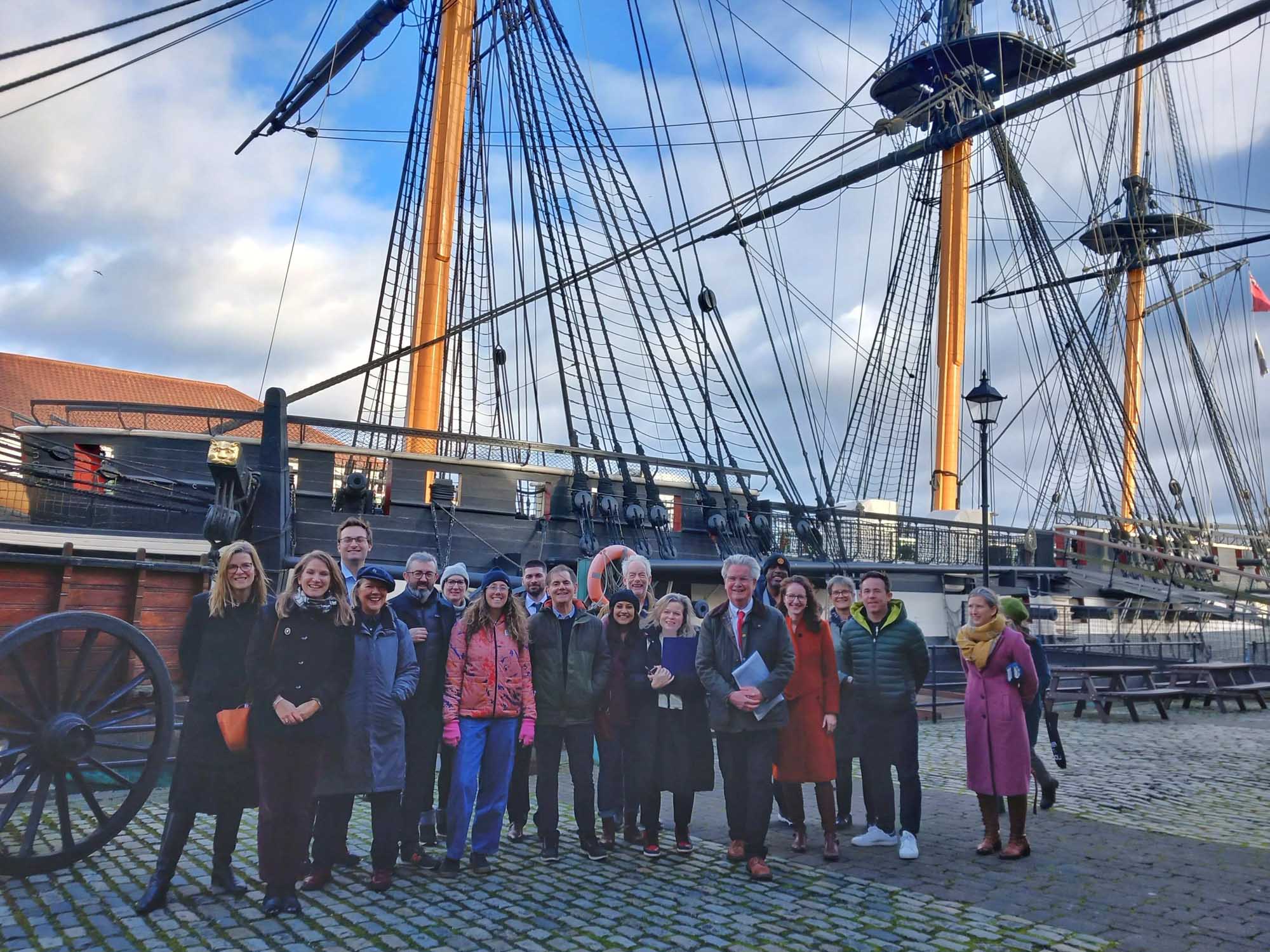 A group of people standing in front of an old sailing boat. 