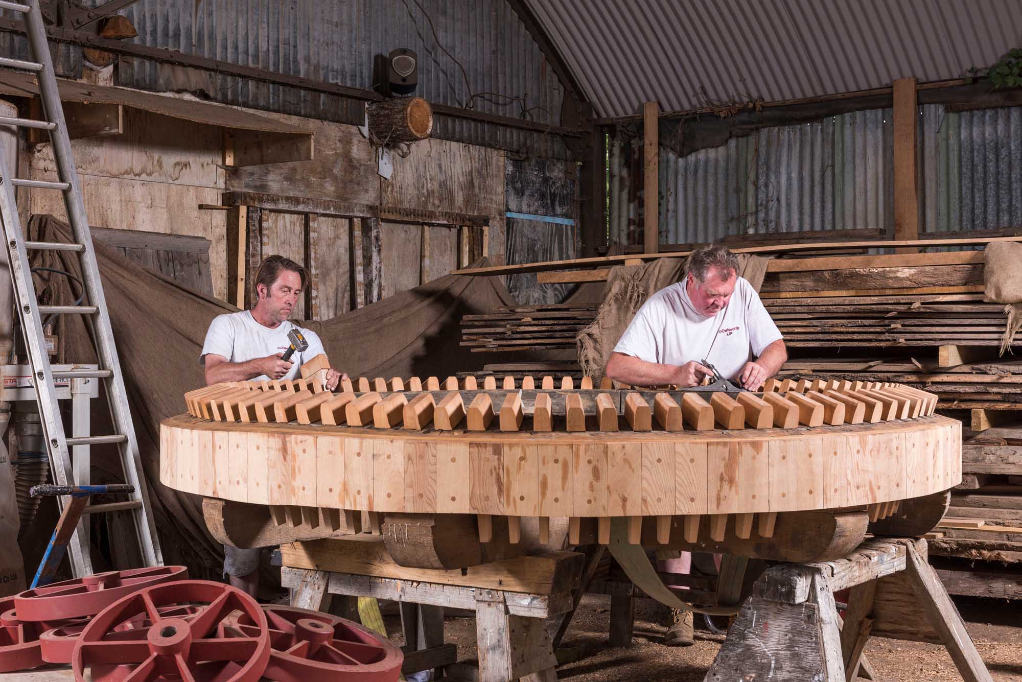 Two men working on a large wooden wheel in a warehouse.