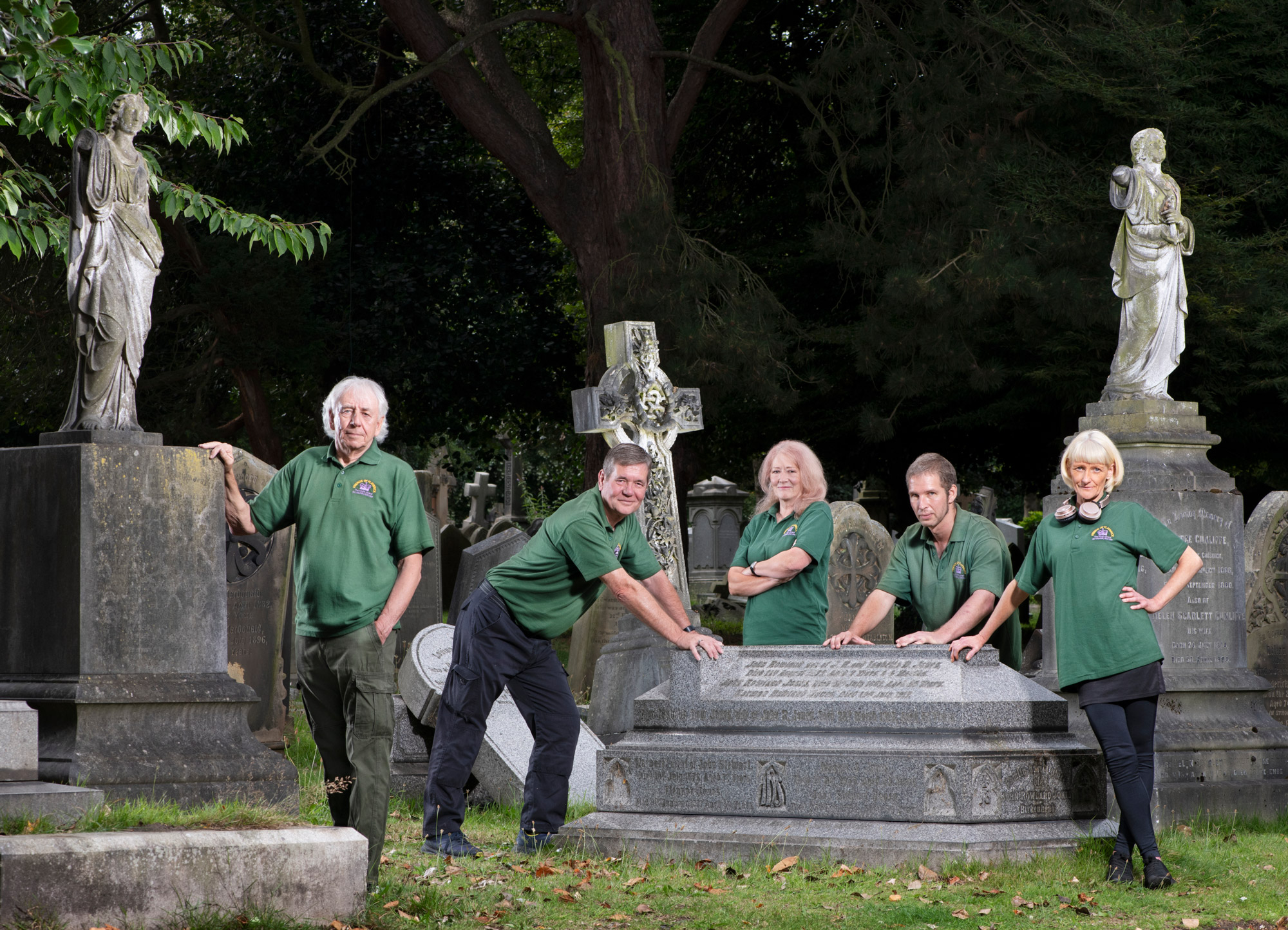 A group of 5 people wearing green polo shirts branded Friends of Flaybrick Memorial Gardens pose for a group photo among the tombstones in a memorial garden.