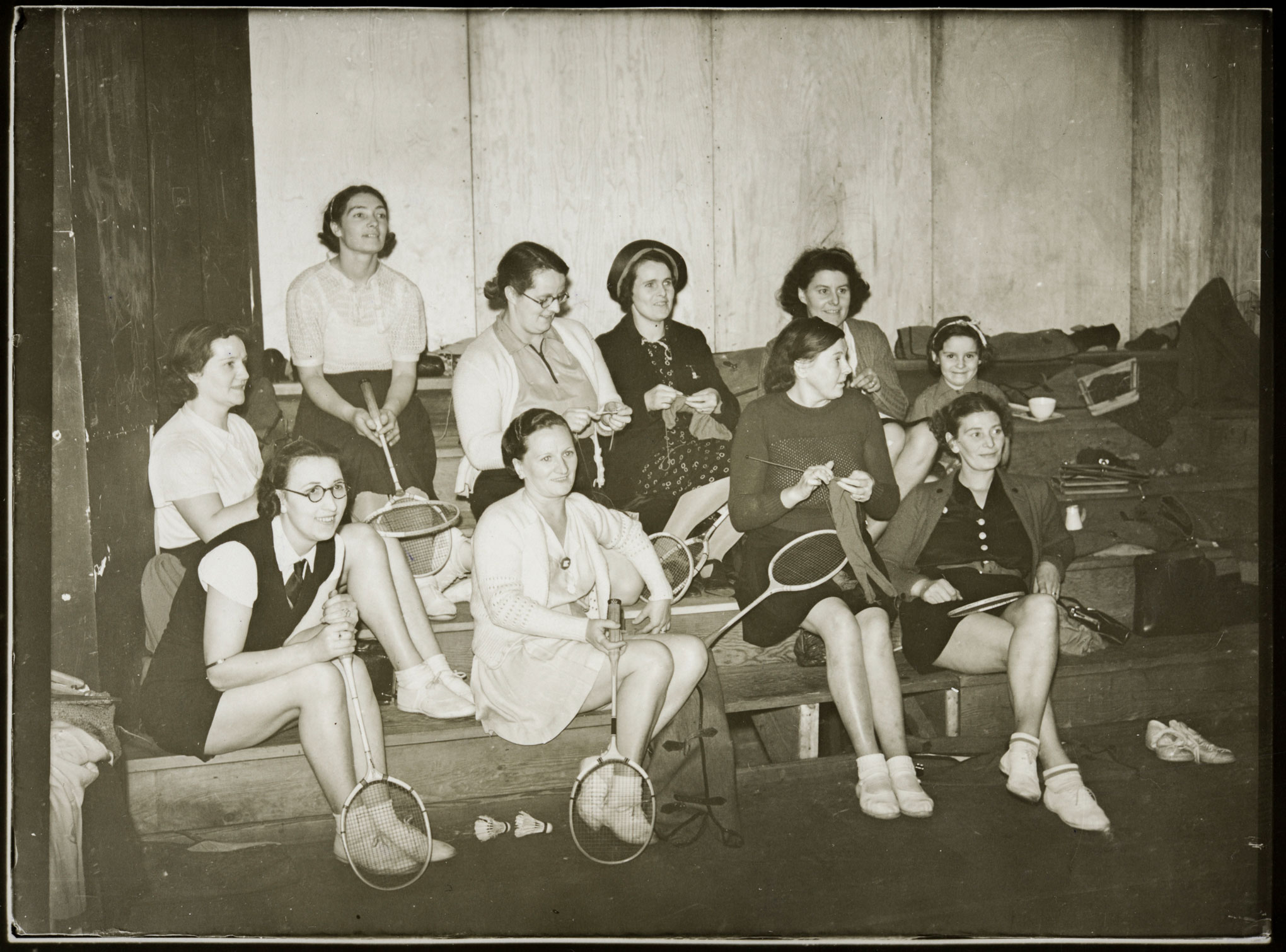 Women sitting on tiered benches holding badminton racquets and knitting.