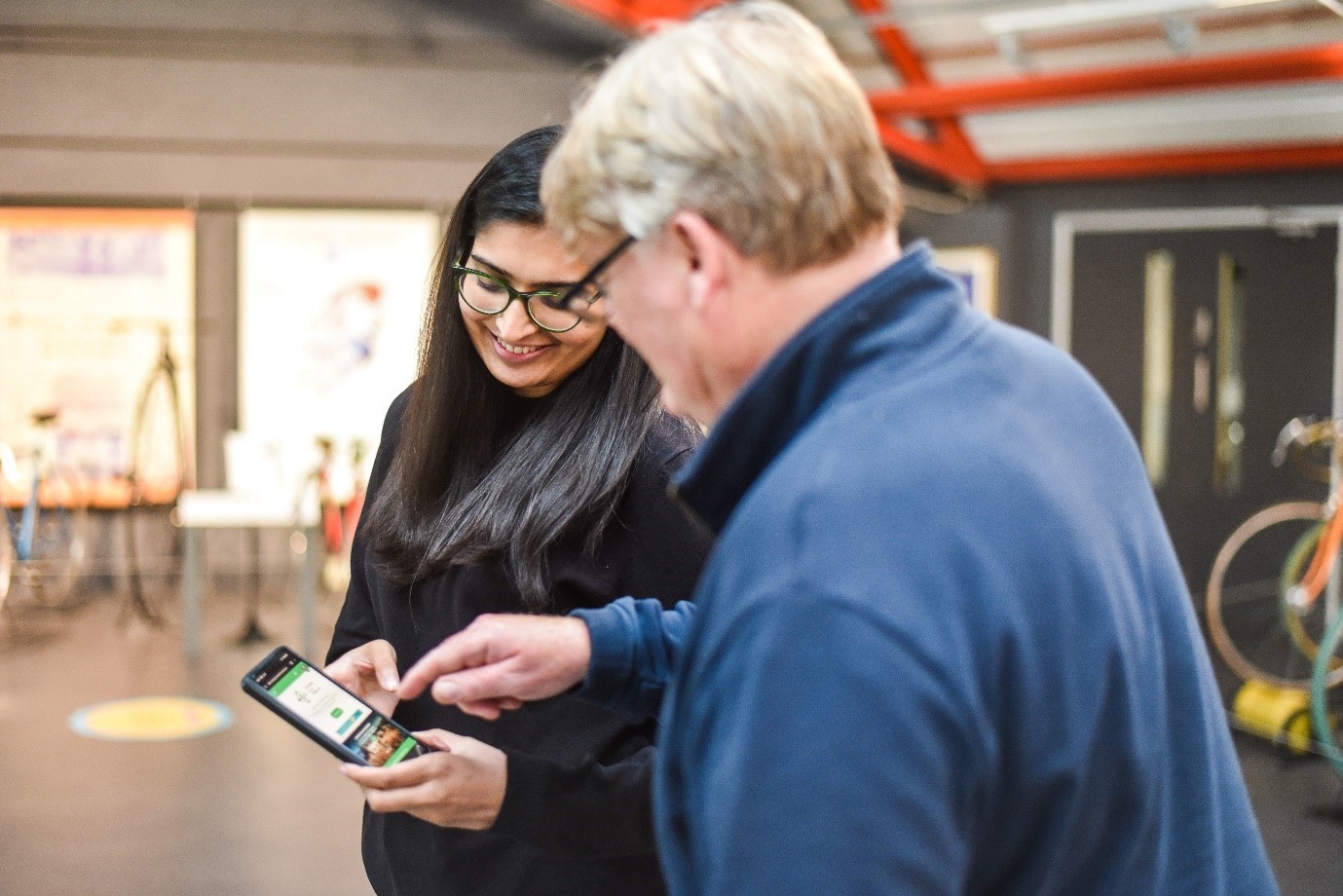 A woman and an older man stand together in a brightly lit indoor space. The woman is holding a smartphone, showing something to the man, who is pointing at the screen