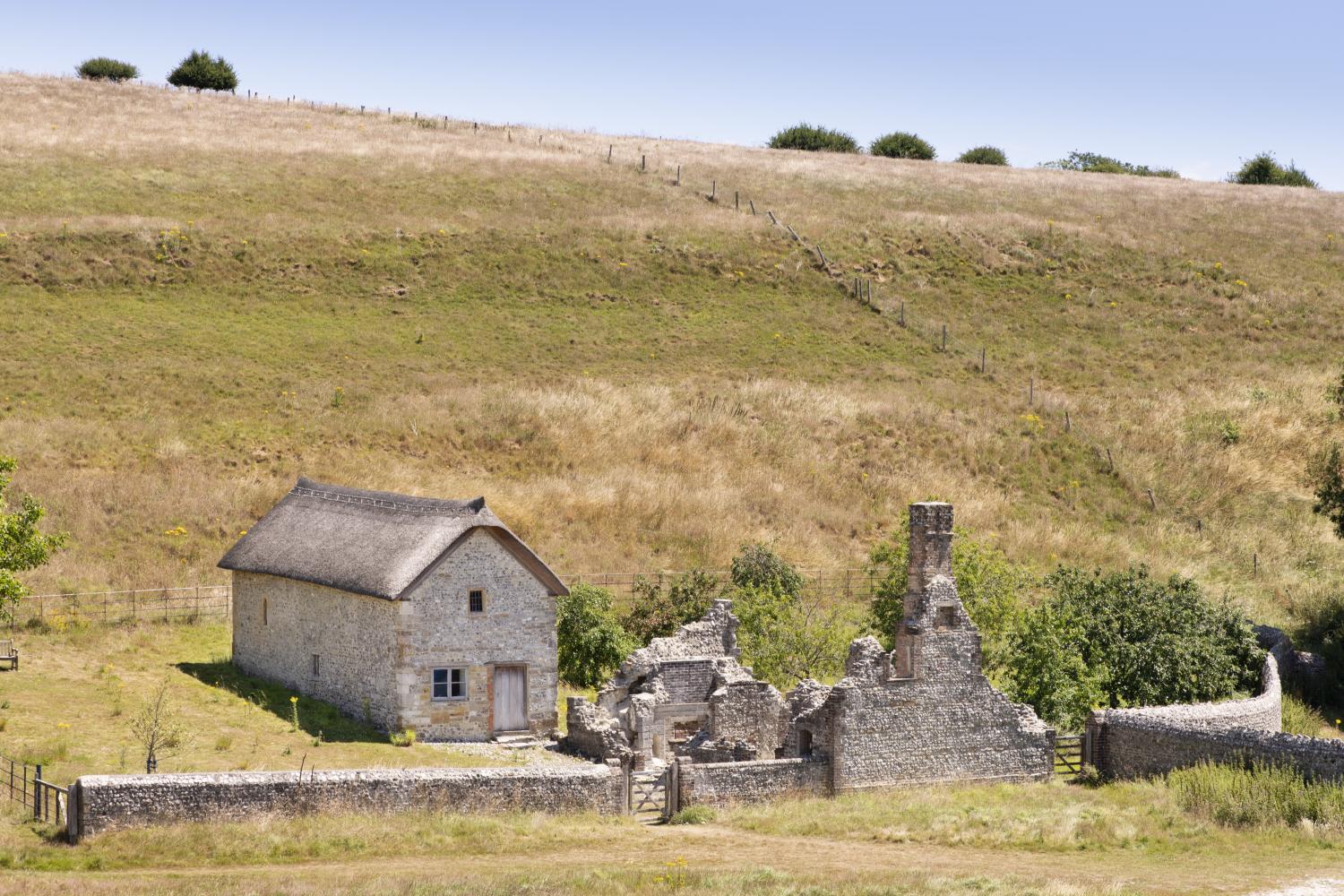 Photograph of a field with two structures in the middle with stone walls stretching out from each side. One structure has no roof and is a ruin, the other has a roof.