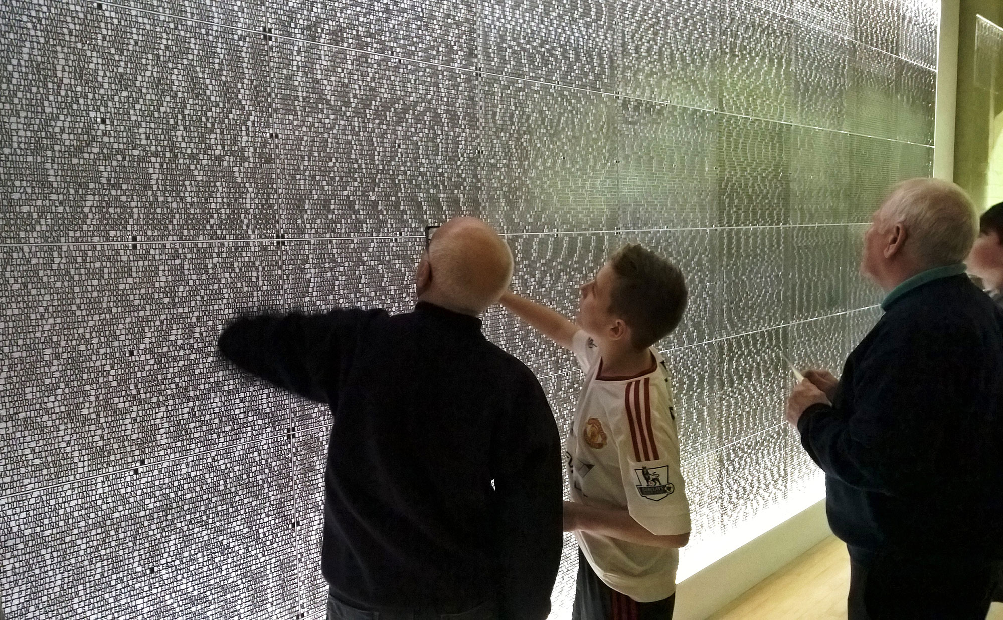 2 older men and a young man examine a wall, covered in black lettering on a white background.