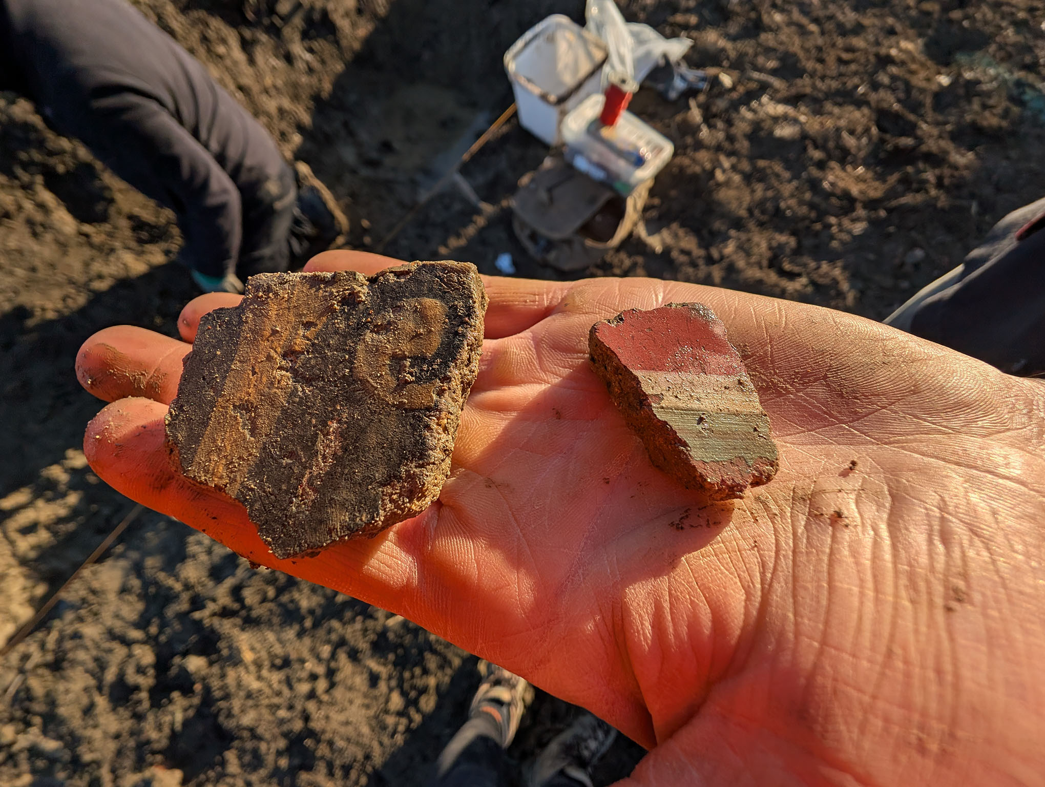 Close up image of a hand holding two pieces of Roman painted plaster. In the background is earth and equipment from an archaeological dig.