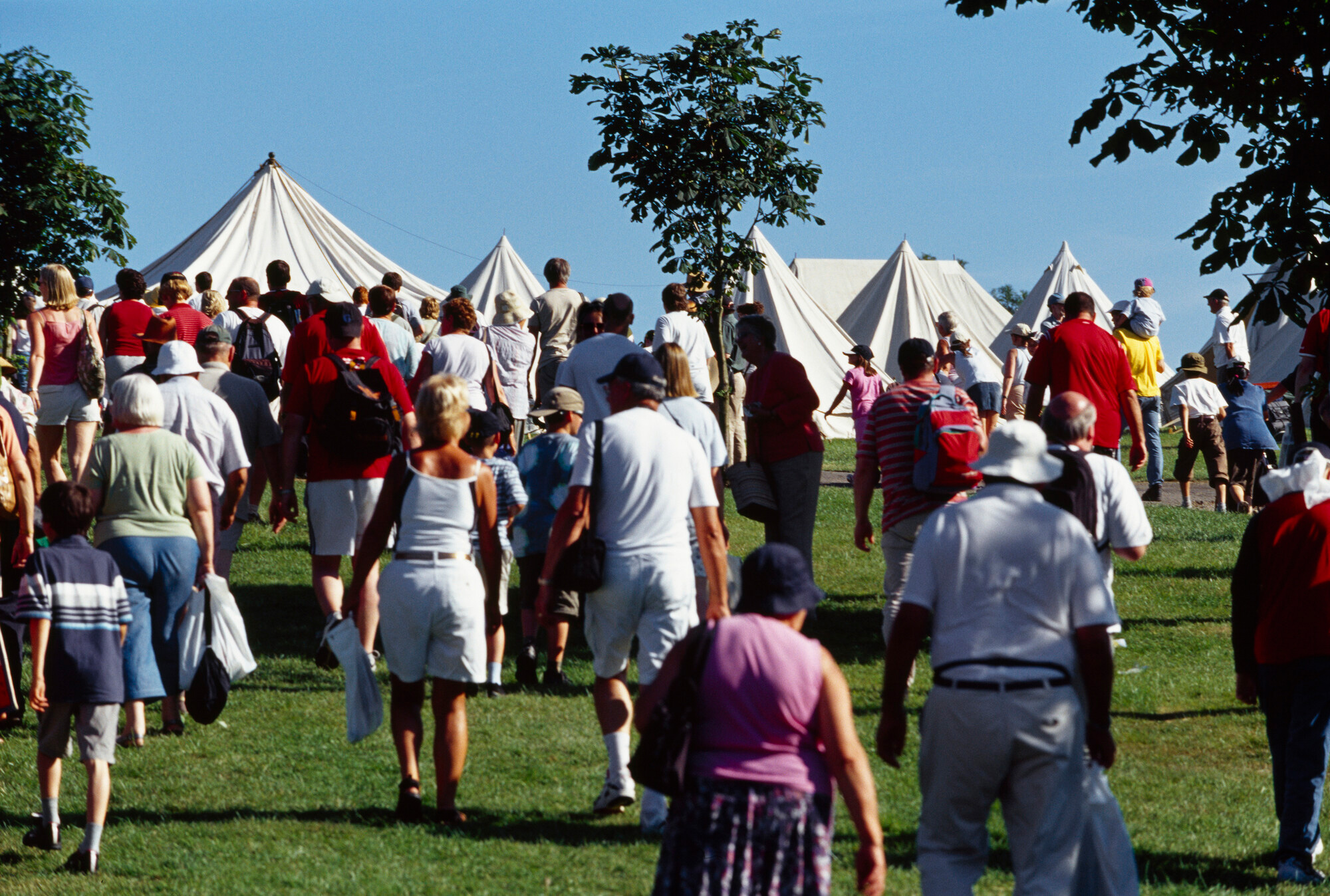 A large crowd of people walks towards white tents on a sunny day, surrounded by green grass and trees, creating a lively outdoor atmosphere.