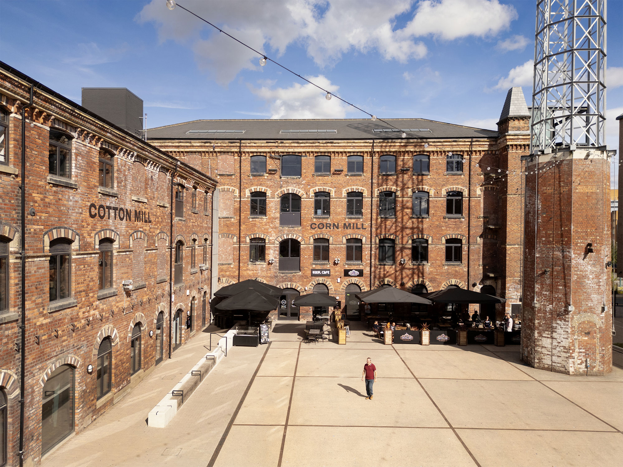 A red brick building with big arched windows wraps around a paved courtyard. Wording on two walls of the building reads Cotton Mill. There are large black umbrellas covering tables for a cafe along one side.