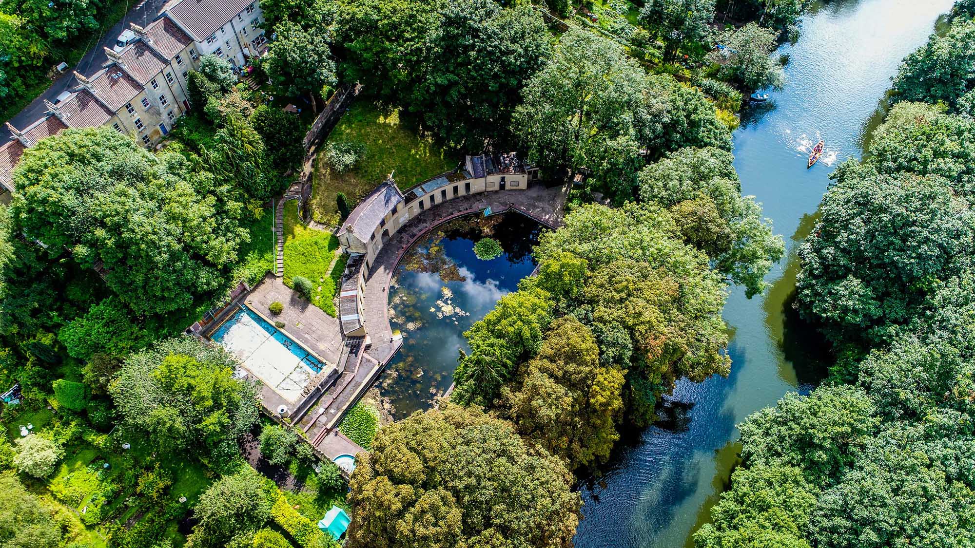 An aerial view of a crescent-shaped outdoor swimming pool set beside a river surrounded by trees and gardens