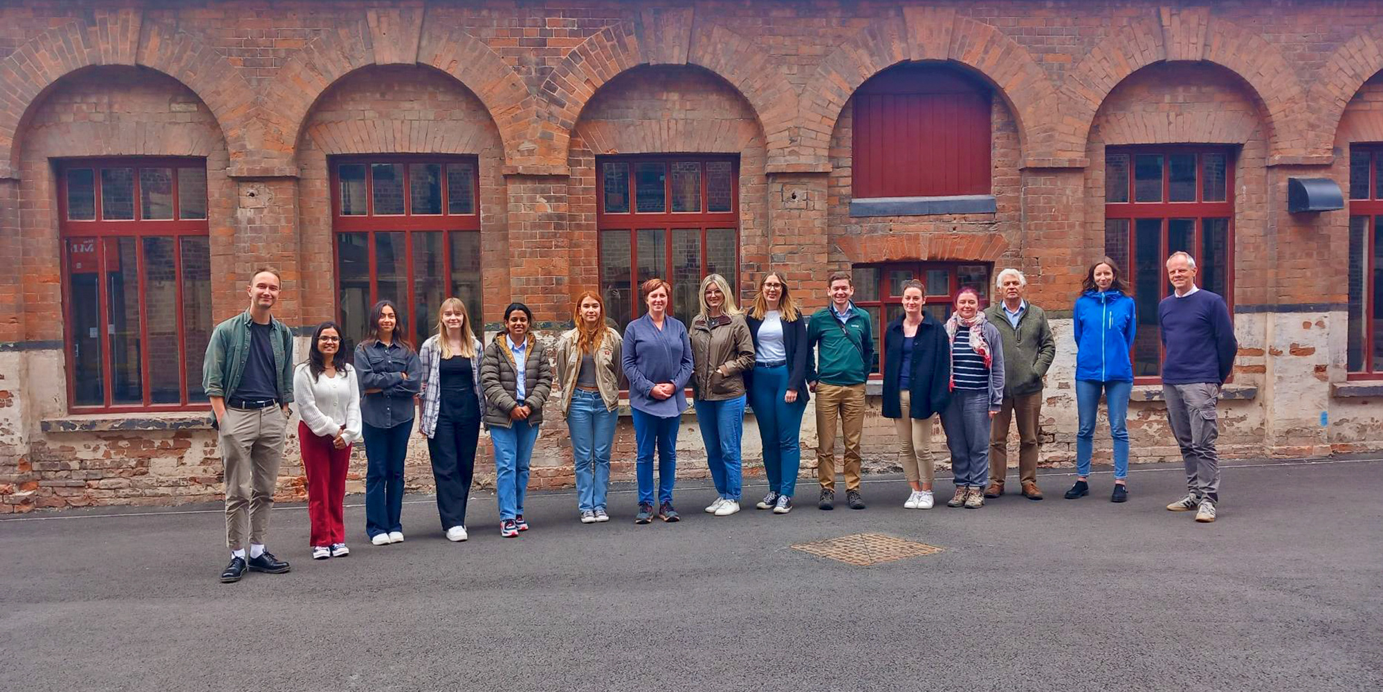 A group of 15 individuals lined up in front of a red brick industrial building. 