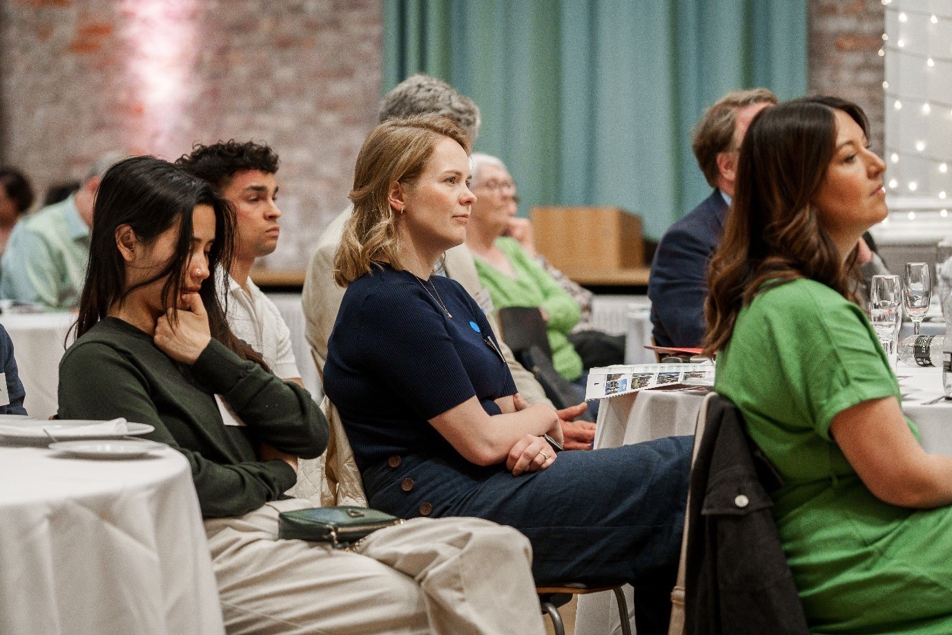 A group of people attending a conference sit at round tables, attentively listening to a speaker. 