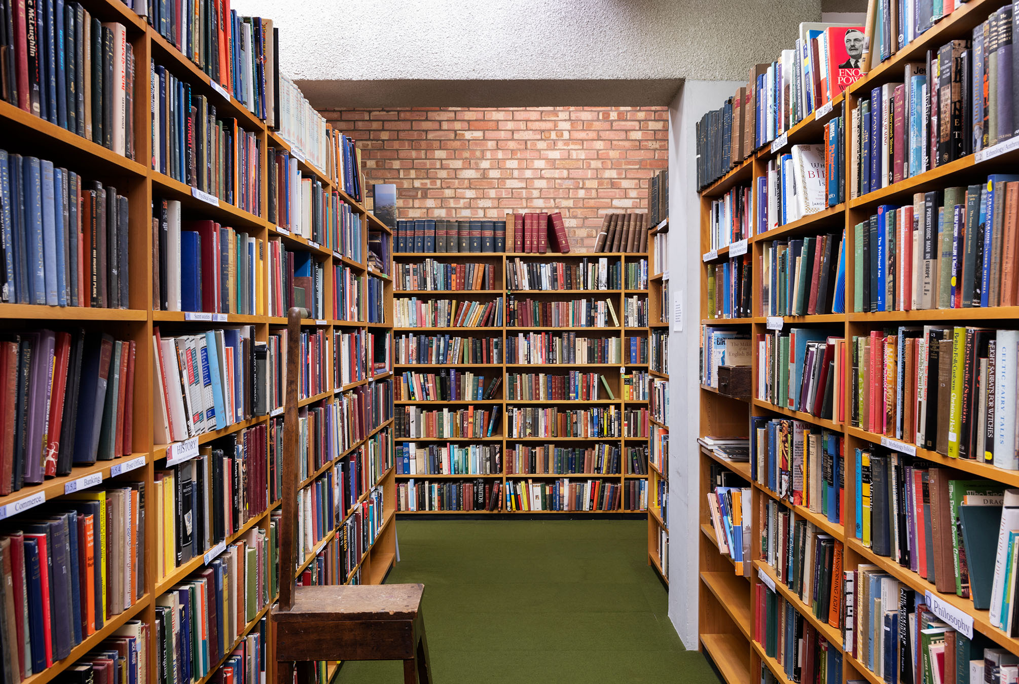 A photograph of a library interior with shelves filled with books, a green carpet and a red brick wall in the background