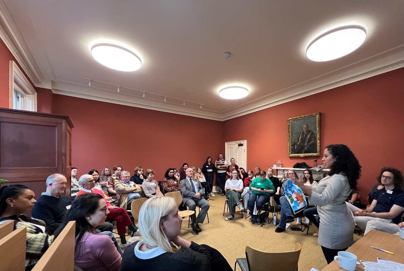 A woman stands at the front of a room, addressing a seated audience with a colorful booklet in her hand. The room has bright walls, modern ceiling lights, and a portrait painting on one wall