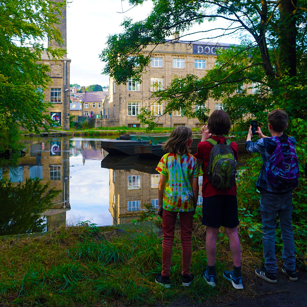 Three children stand on a canal bank, photographing the canal and industrial landscape through overhanging trees.