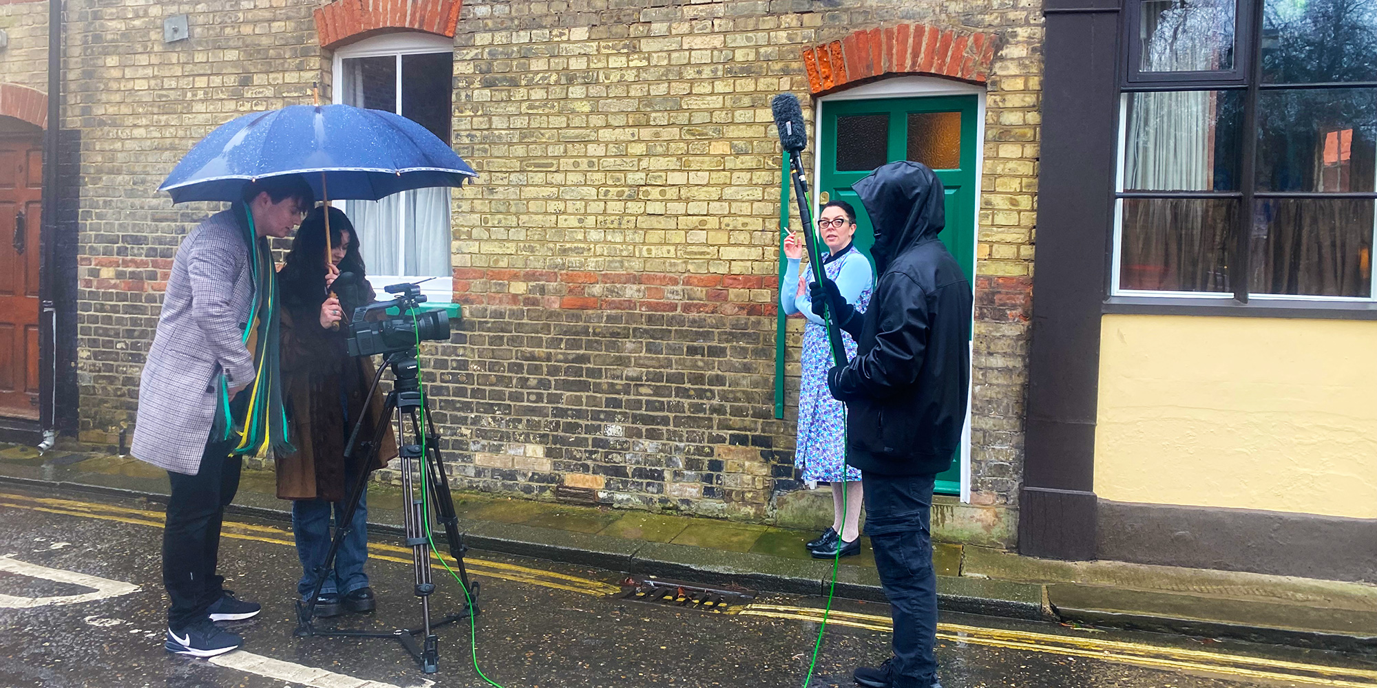 A woman in mid-century costume smokes a cigarette outside a brick townhouse while a film camera crew and microphone operator stand by.