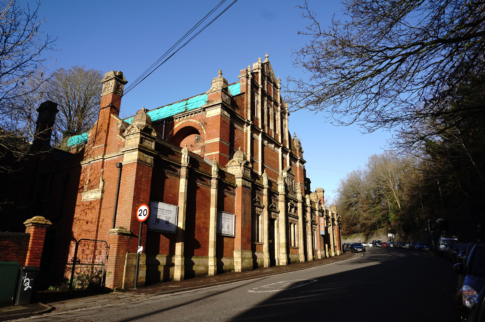 A photograph of the front of a large red brick building with ornate terracotta details facing onto an urban street. The roof has a temporary covering and the building is in poor repair.