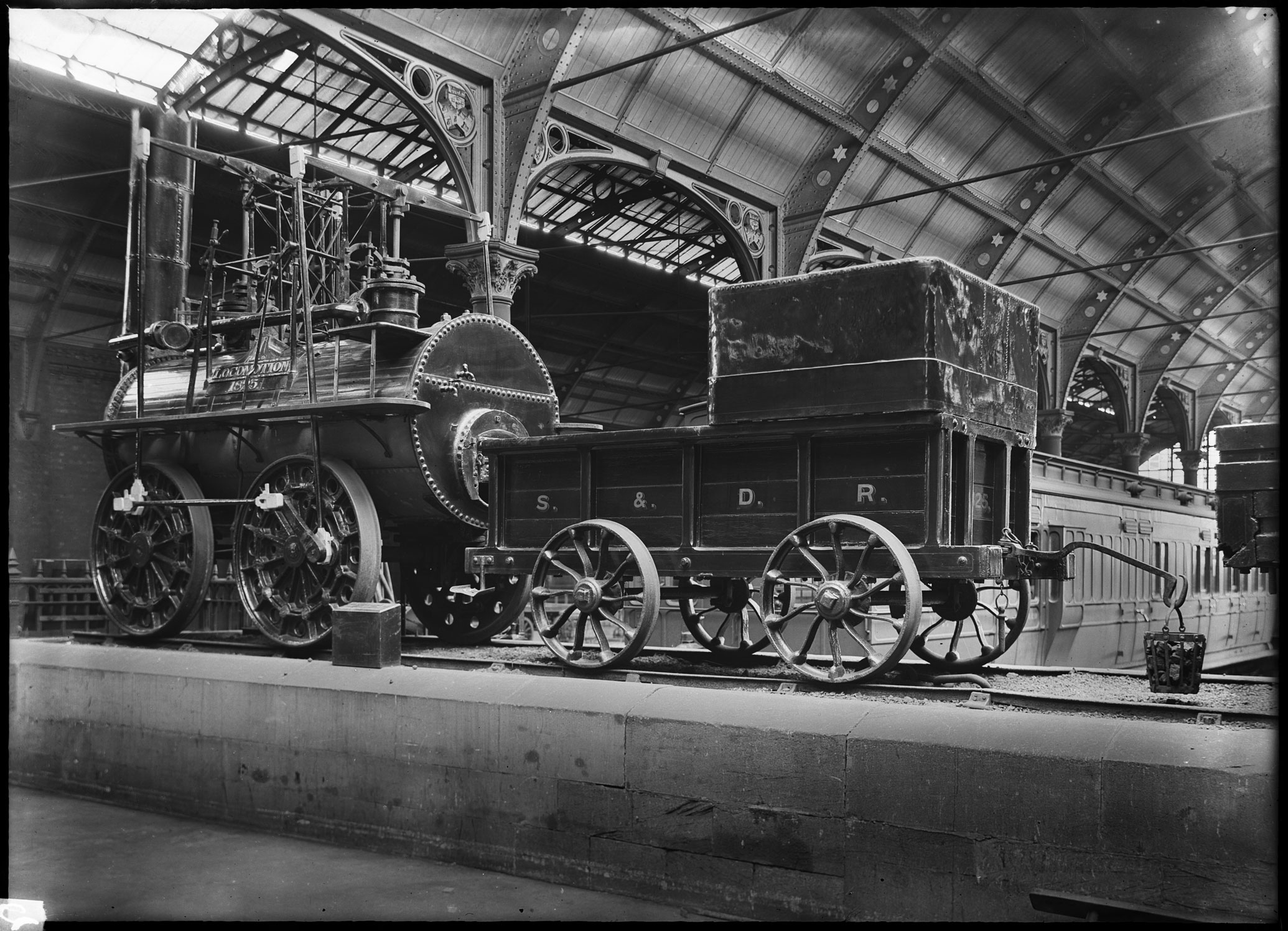 A black and white archive image of an early railway locomotive on display at a railway station.