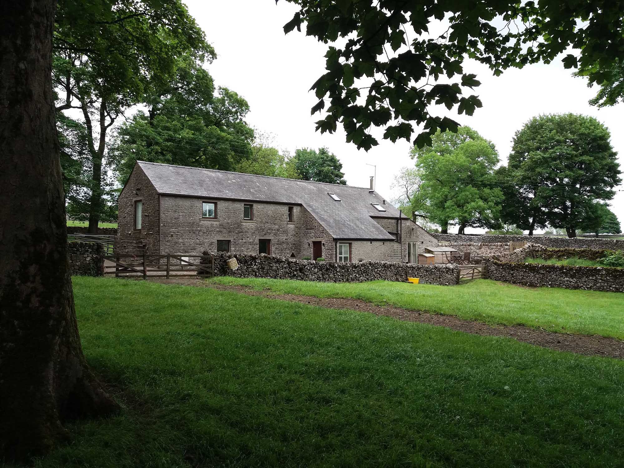 Stone farm building with grey slate tile roof set in fields bordered by dry stone walls with large trees dotted around and a dry stone wall running in front of the house.