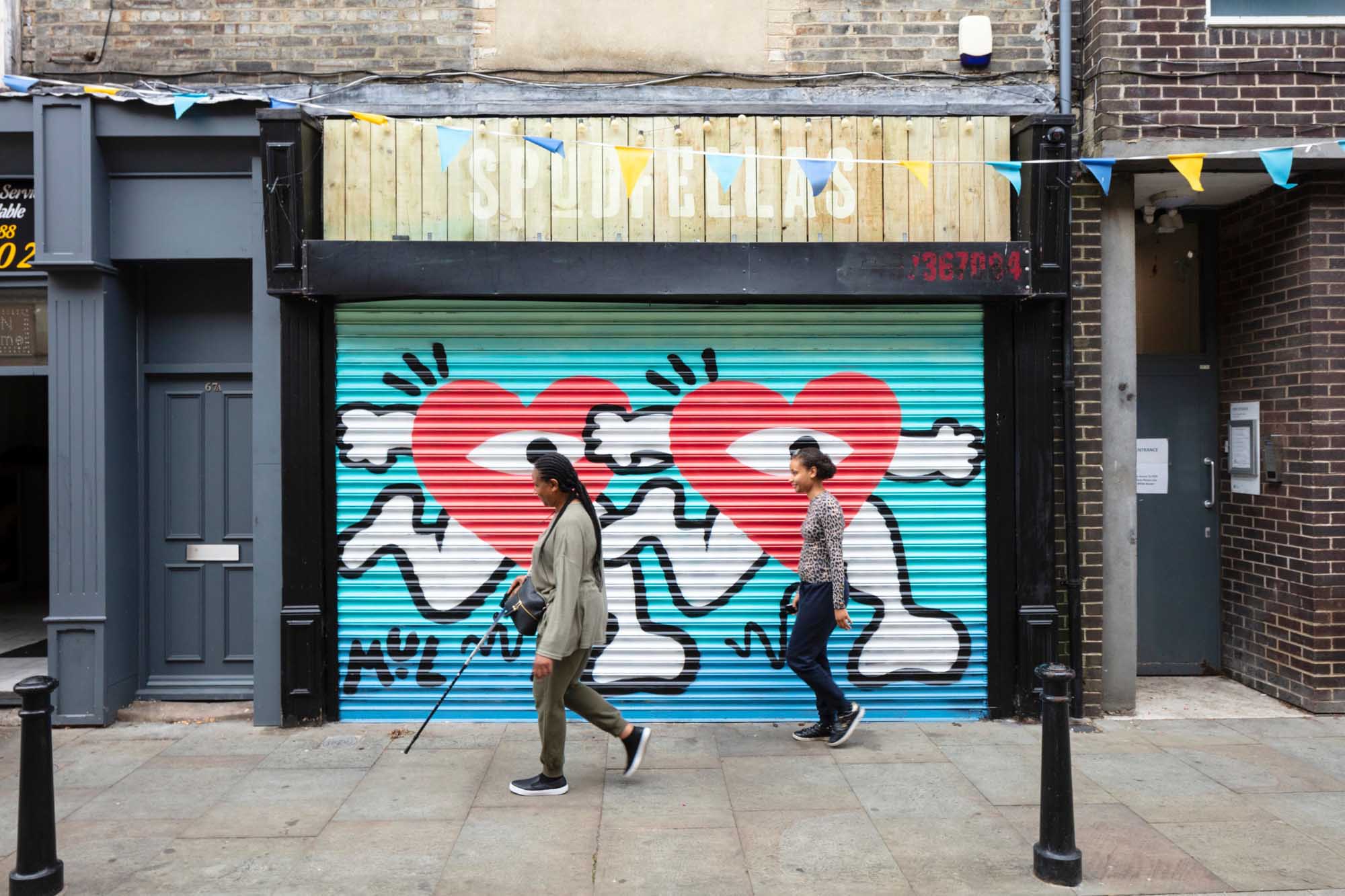 A street scene of a woman with a cane and a young person walking past a shop front.