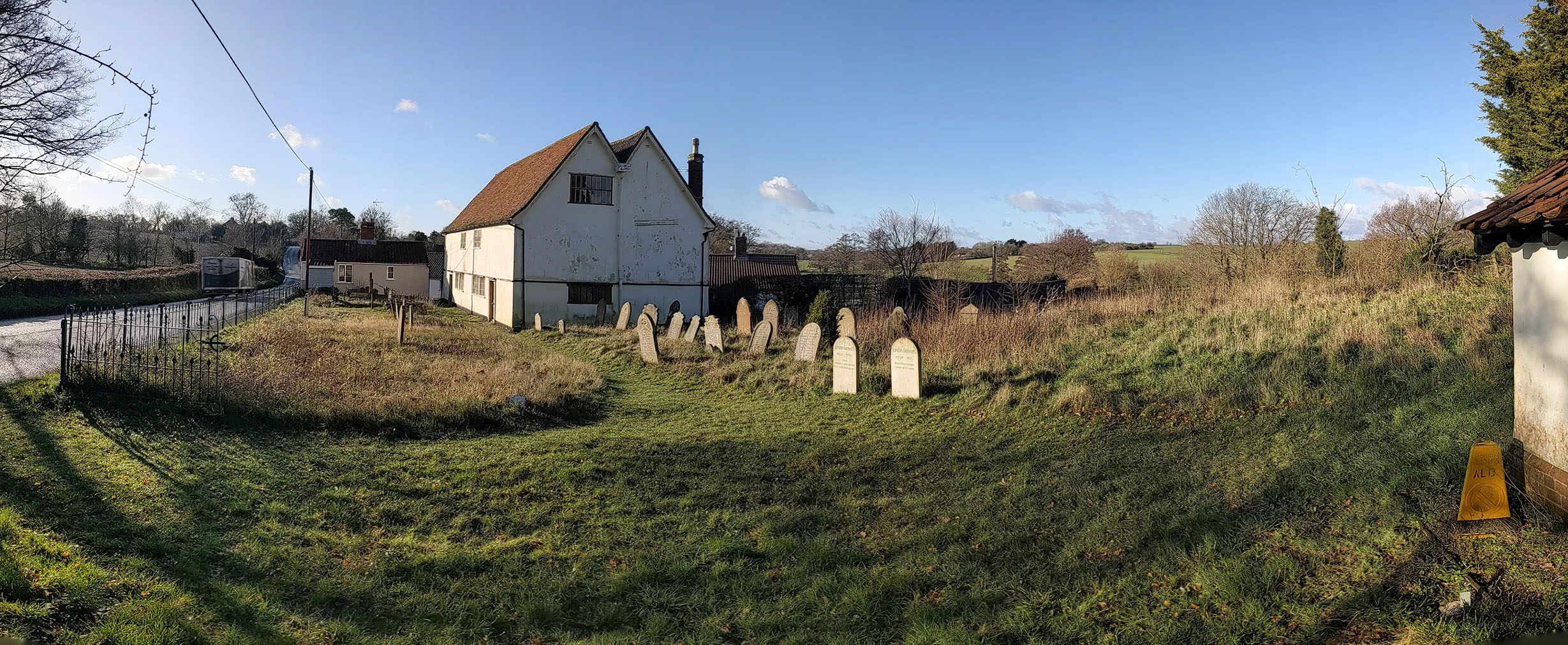 The photograph shows a white building with double roof in brown tiling, with gravestones dotted around and a scrub landscape