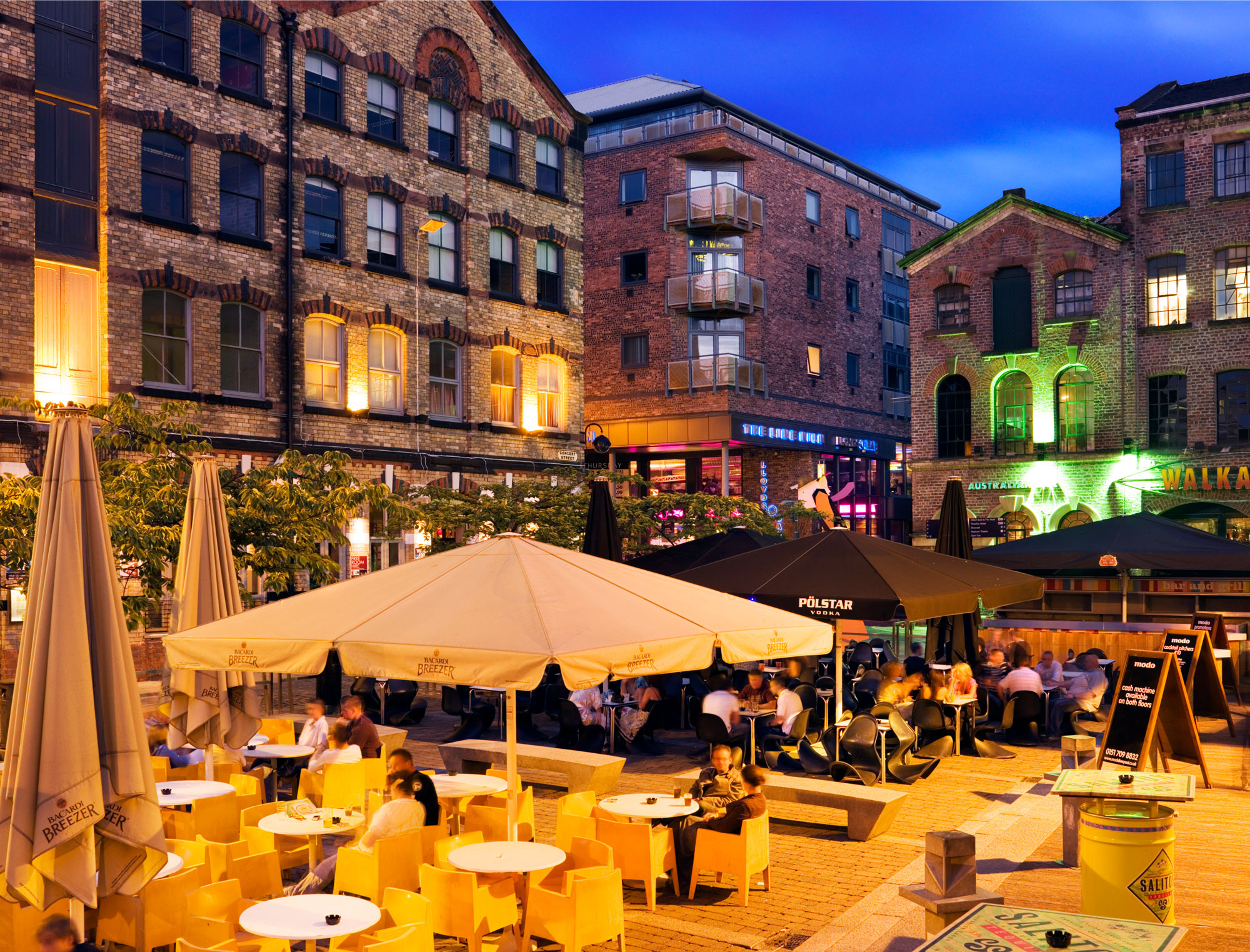 Colour photo taken at dusk showing a square where people are socialising at tables under parasols. Surrounding the square are old industrial buildings that have been restored and are in places lit up.