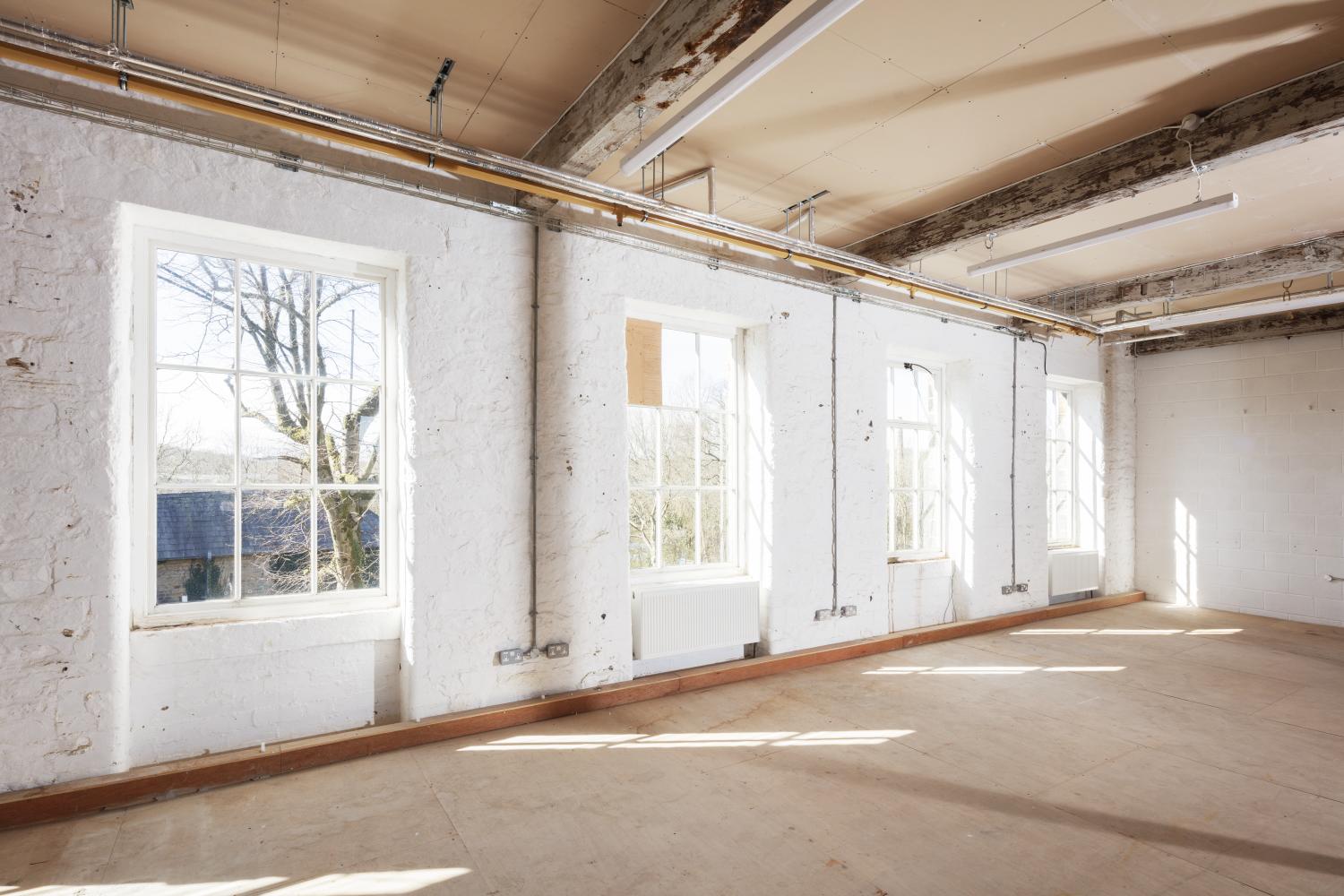 Internal view of a whitewashed wall with four large windows, the floor and ceiling are unfinished plywood.