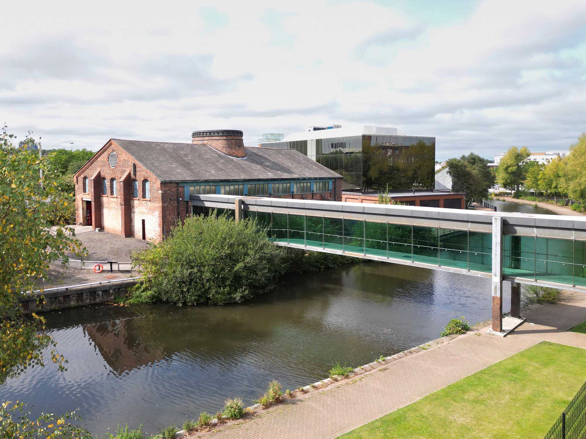 A glass-enclosed walkway crossed a canal to reach a red brick building, with a modern glass building behind.