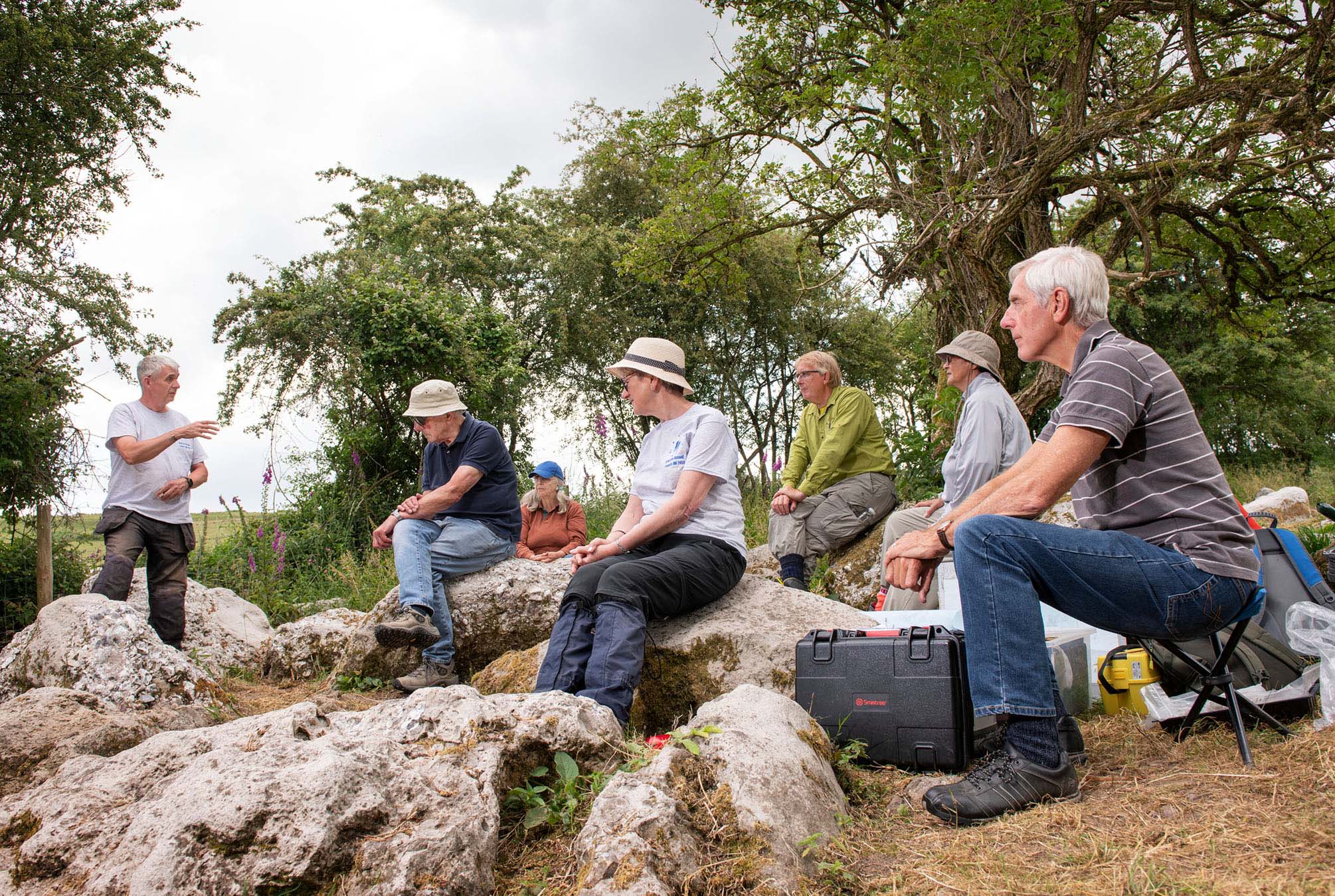 General view of a mixed group of people sitting on rocks listening to a man standing up giving a talk, with trees in the background.