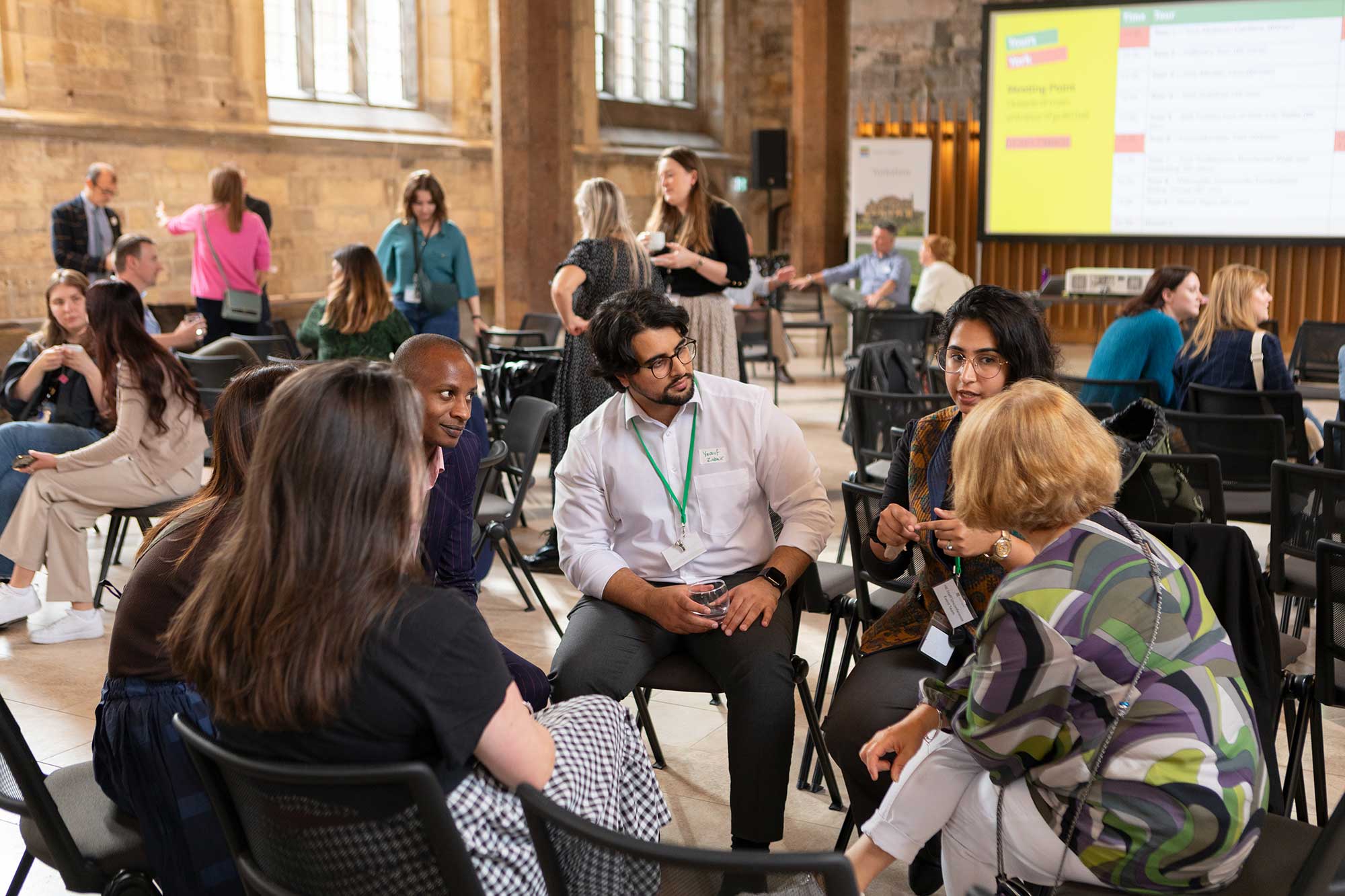 A group of colleagues of varying ages, genders and ethnicities listen to one another talk in an open forum session. Other groups are meeting in the background, and some colleagues are chatting separately. 