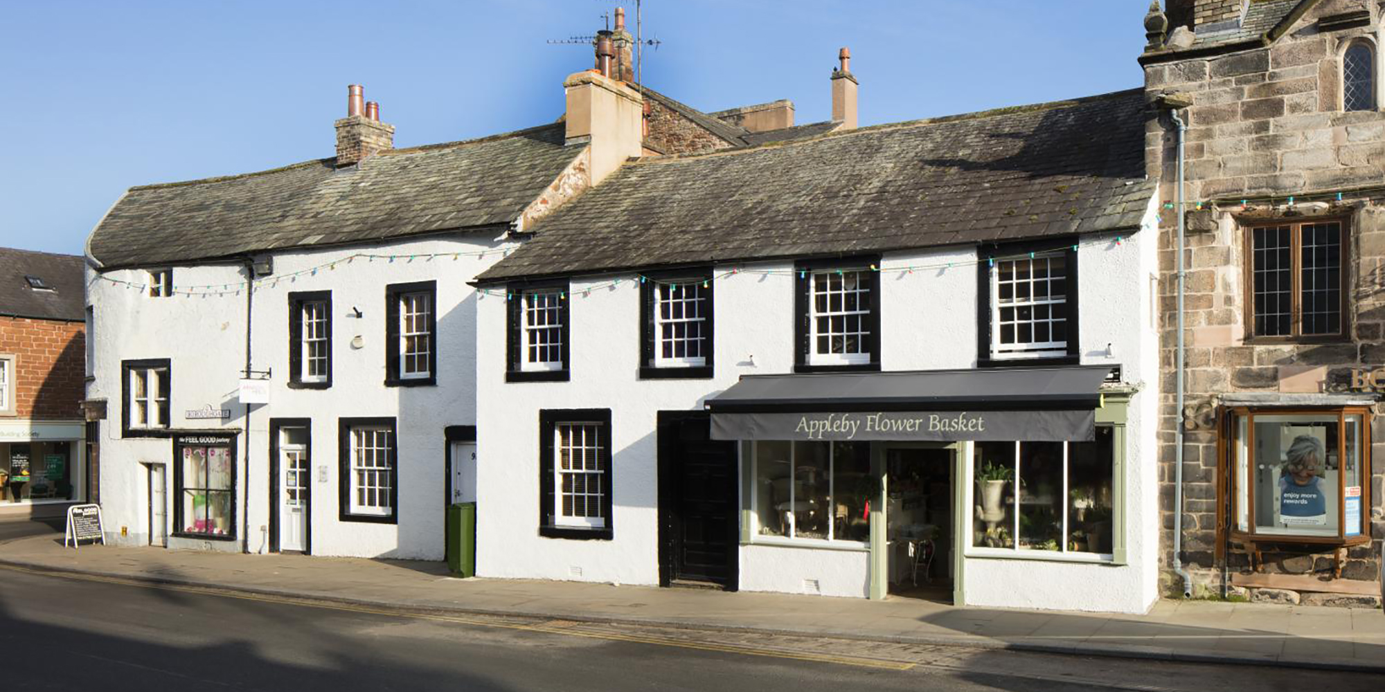 A street-level photo of a street scene focusing on two white brick buildings with black borders around white windows. An awning over the front door of the right hand building has text reading: "Appleby Flower Basket"