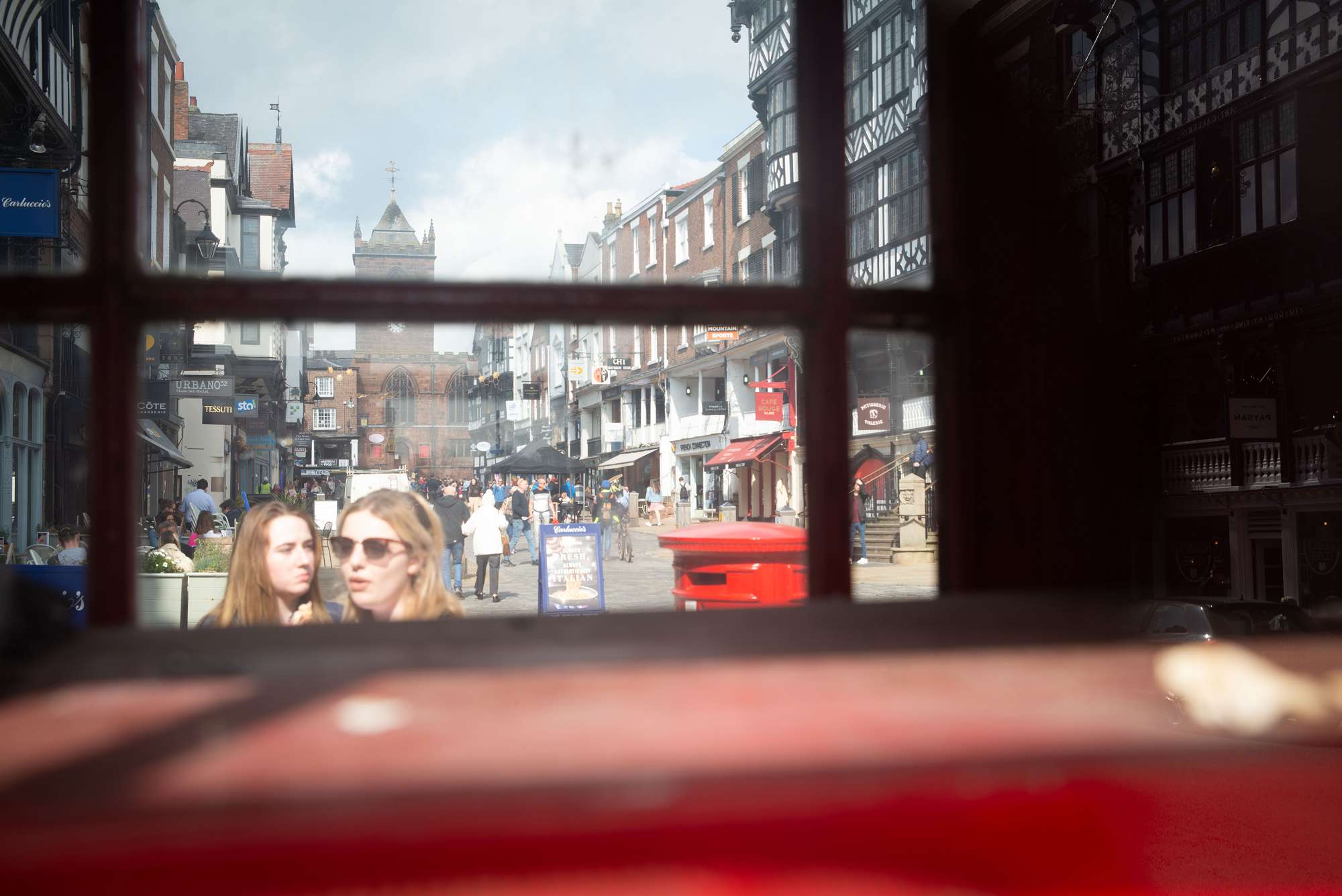 A photo busy historic high street scene is composed through a silhouetted timber-framed window. You can see traditional handing signs, black and white buildings and a church at the far end of the high street.