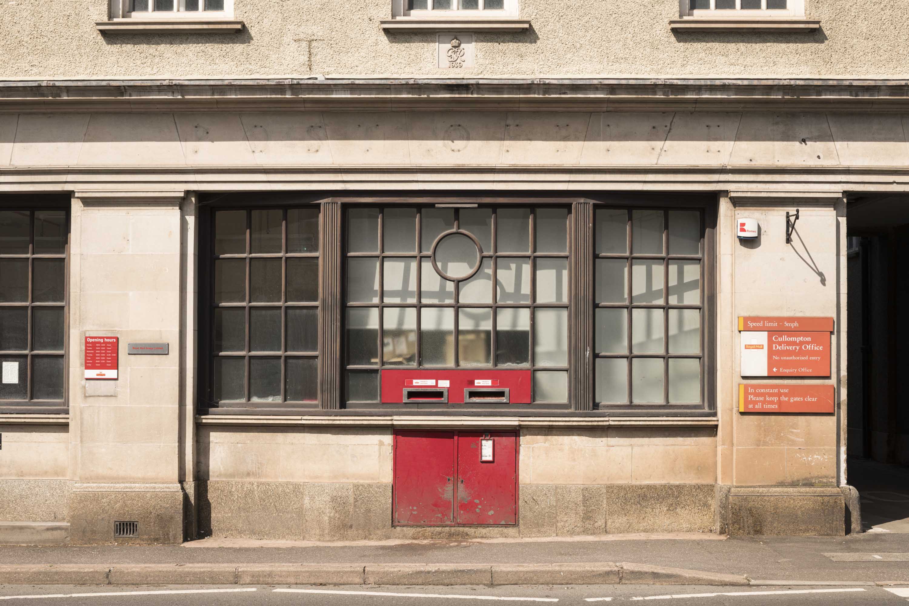 The front of a post office building with a large, multi-paned window above post-boxes which have been set into the masonry.
