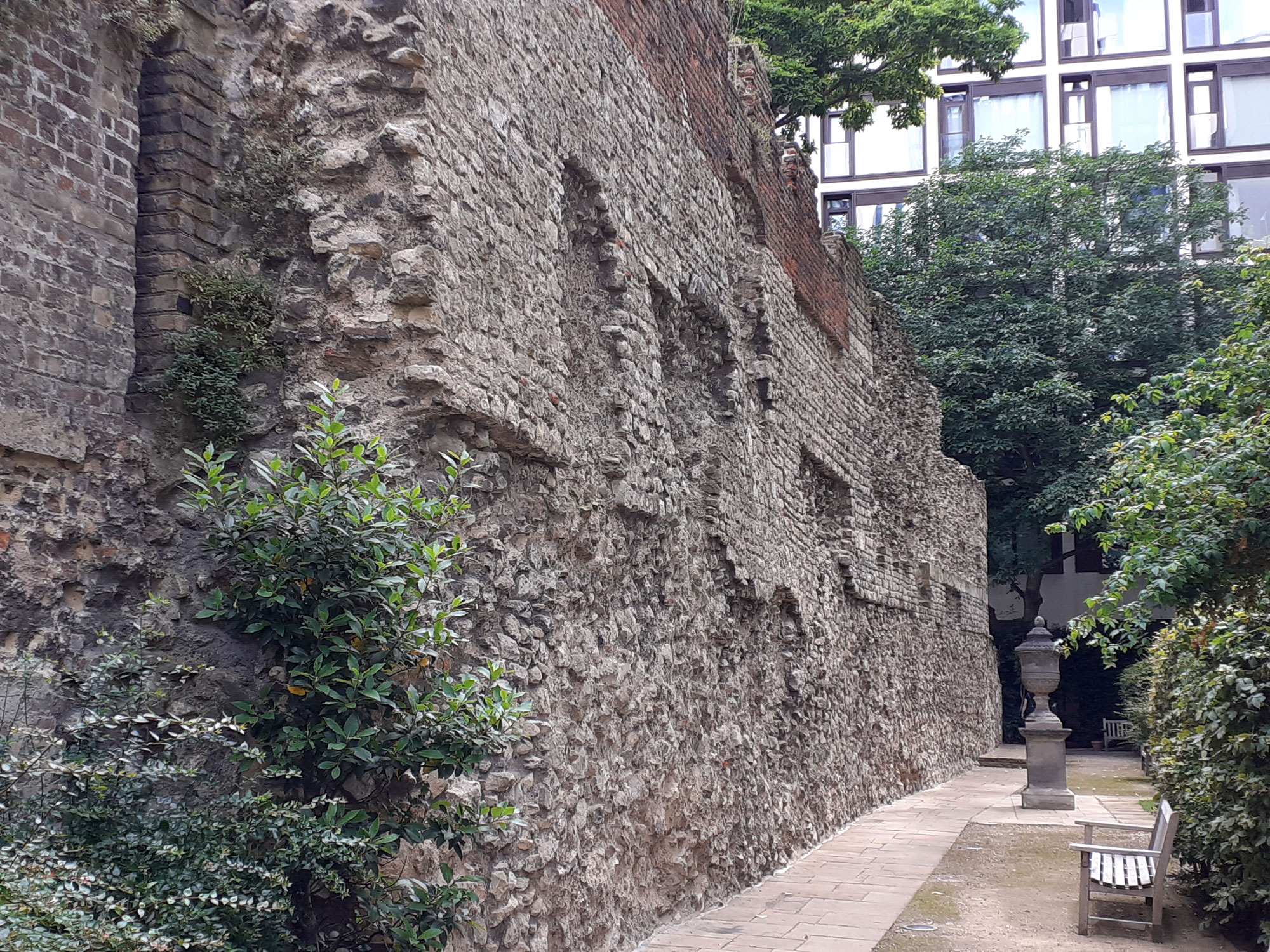 A long old stone wall with some brick sections. In the background, part of a modern building behind it is showing between tree canopies.