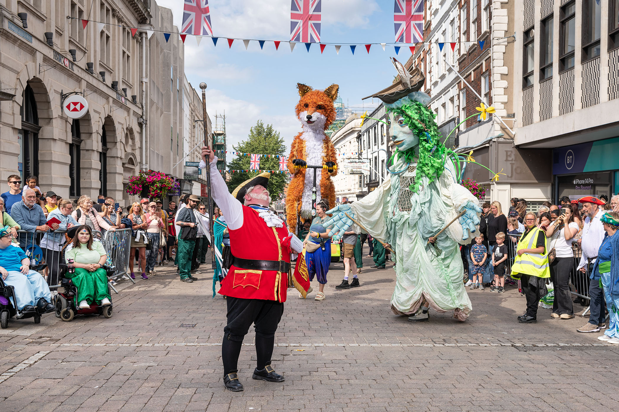 A man in a Lord Mayors outfit stands in the centre of a high street, raising a cane into the air. Behind him are 2 large oversized puppets (one of a fox, the other of a sea maiden). 