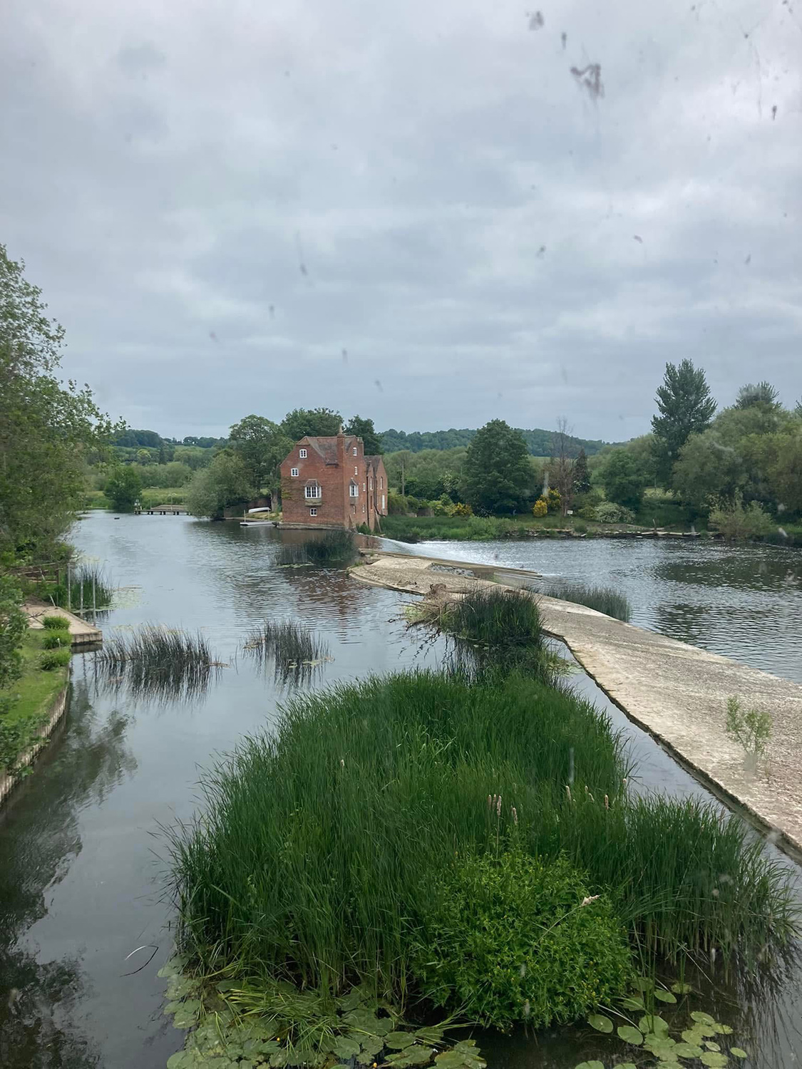 A large expanse of water with reeds in the foreground a red brick house in the background.