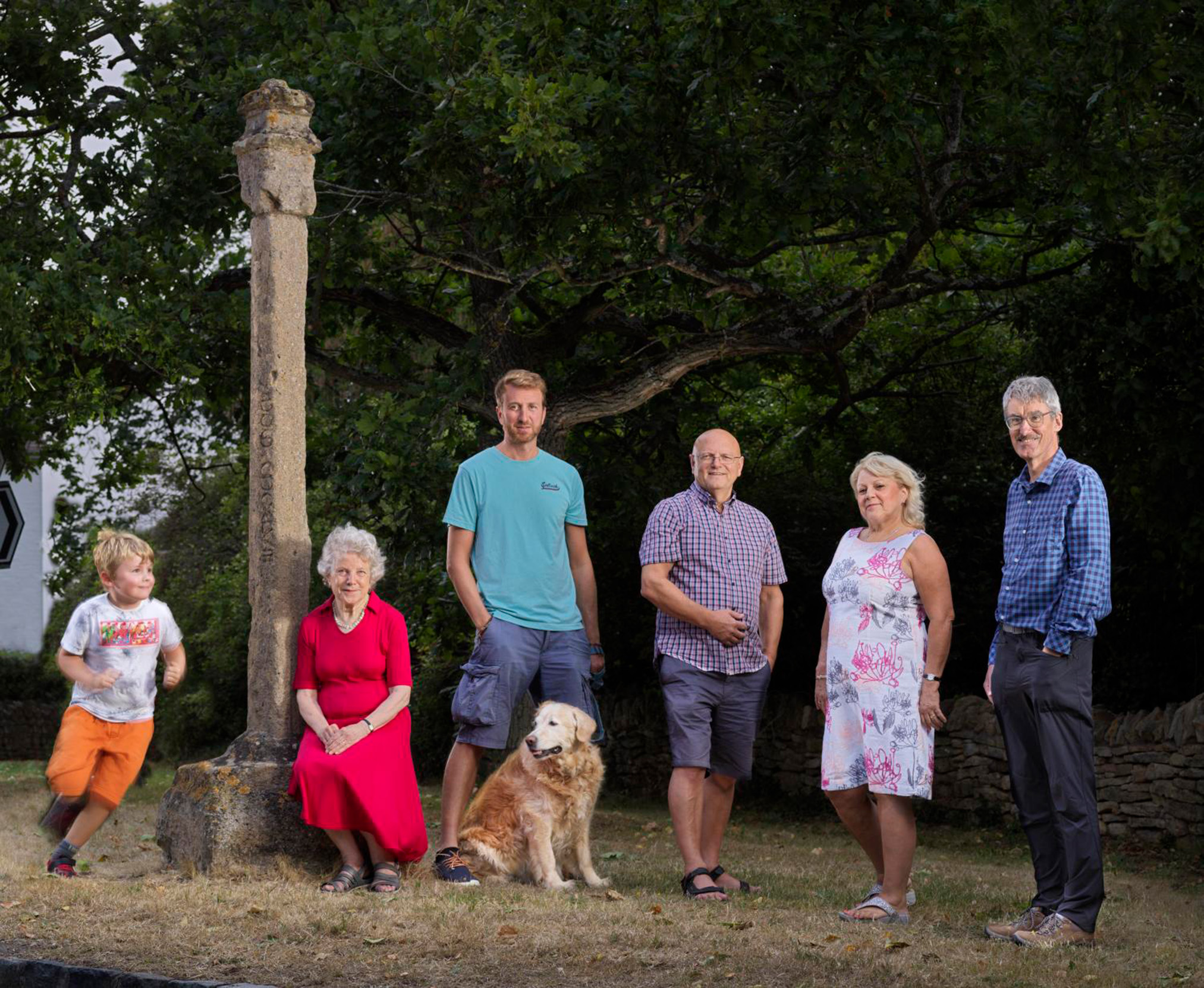 A multi-generational group and a dog posing next to a stone pillar in a park.