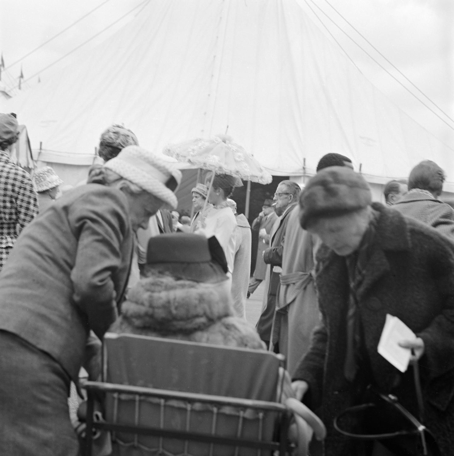 Black and white photo of people outside an exhibition tent. A woman in a wheelchair is visible from behind in the foreground.