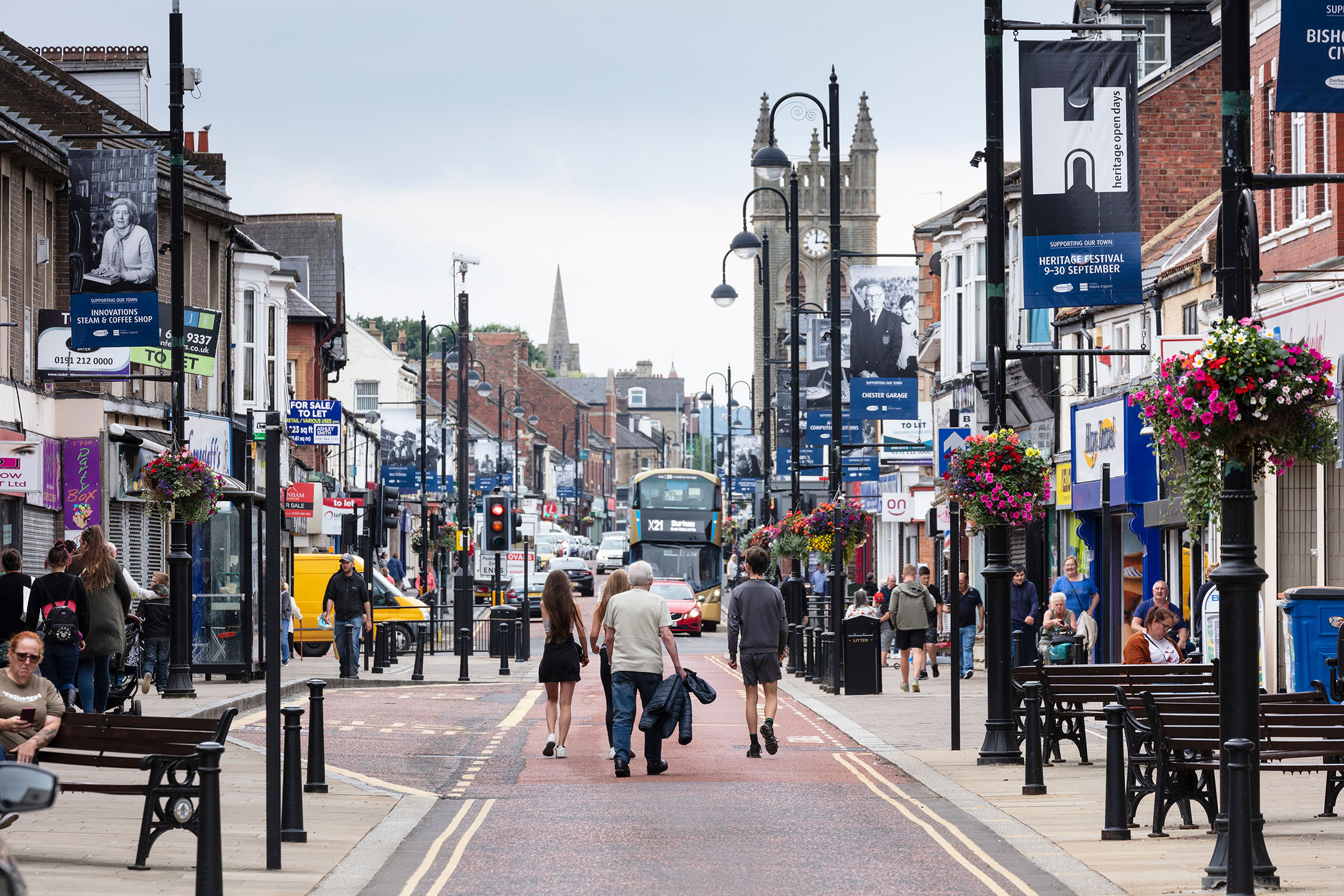 A high street scene featuring people, benches, lamp posts, shops, and traffic in the distance.