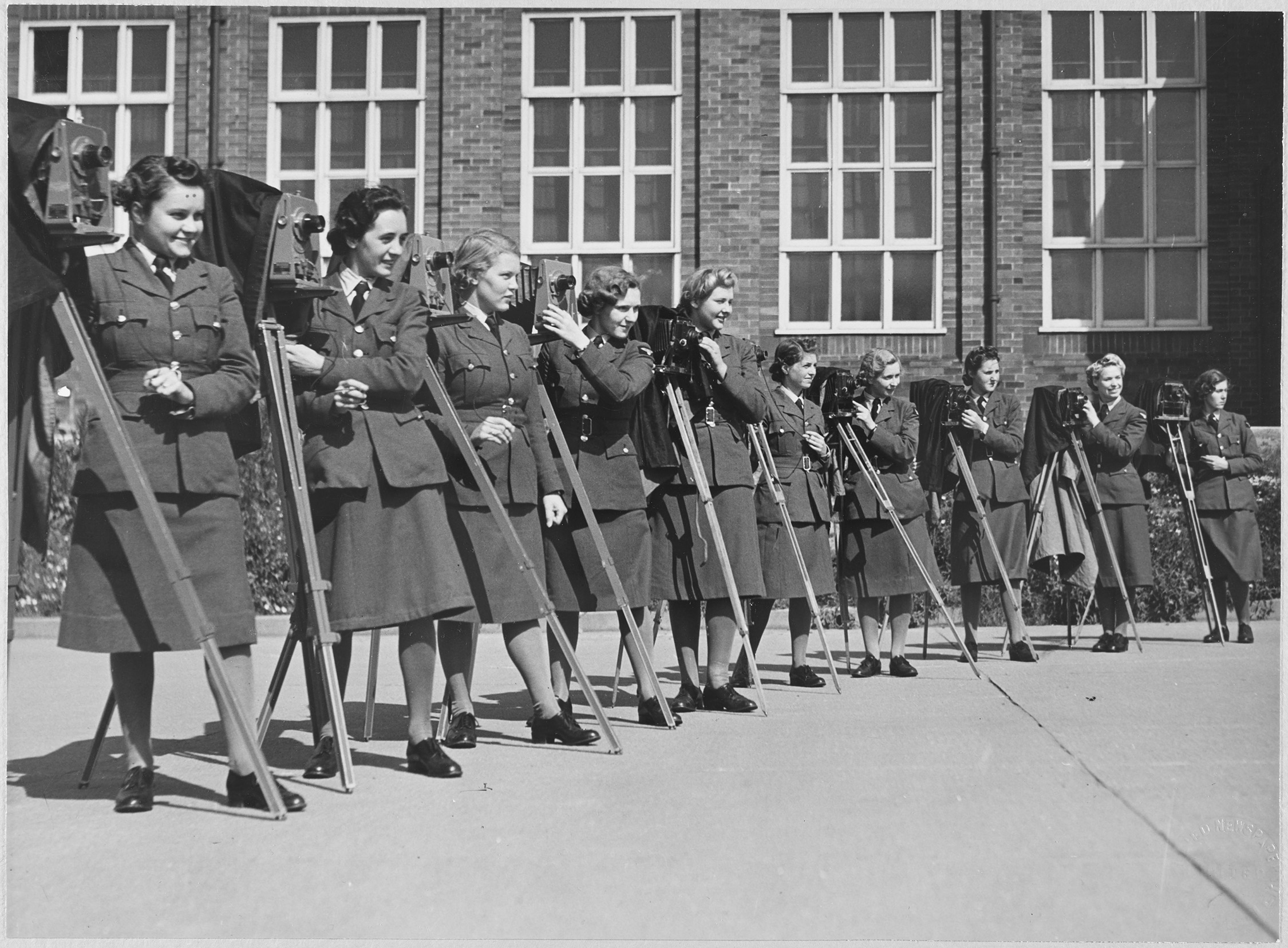 Black and white photograph showing a row of women in military uniform, standing next to view cameras on tripods. They stand in a yard or parade ground. In the background is a brick building with tall windows, three lights wide.