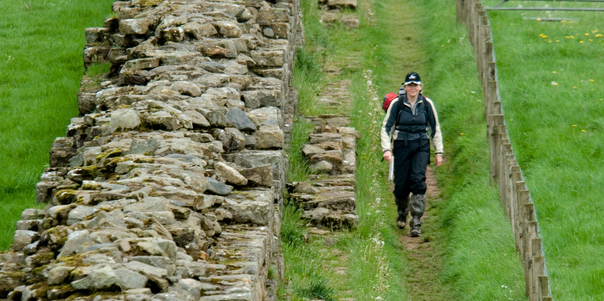 A hiker with a map walks along a trail beside a ruined Roman wall.