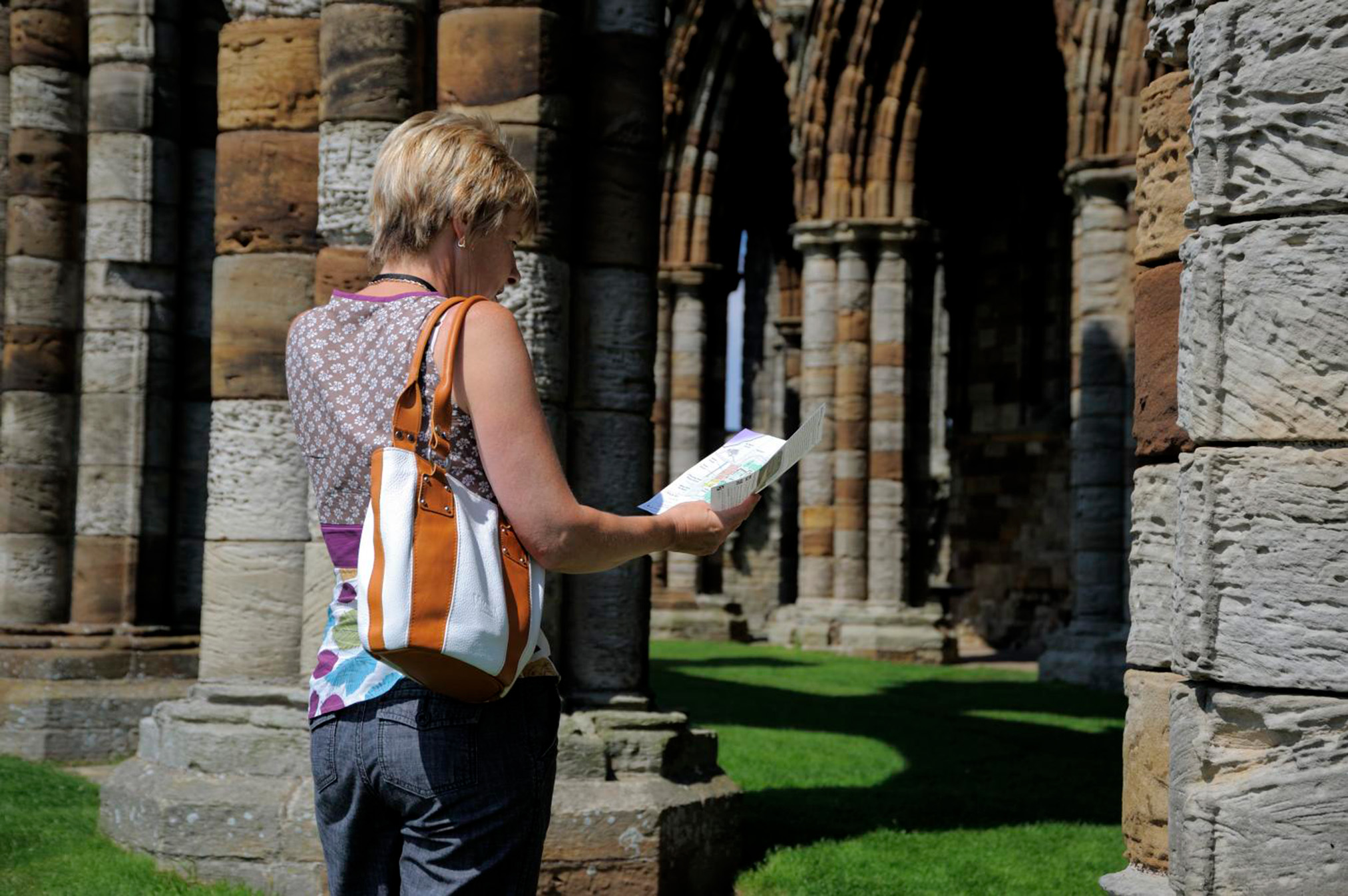 A person with short blonde hair, wearing a bag. Reading a brochure inside the columned archway of an ancient stone building