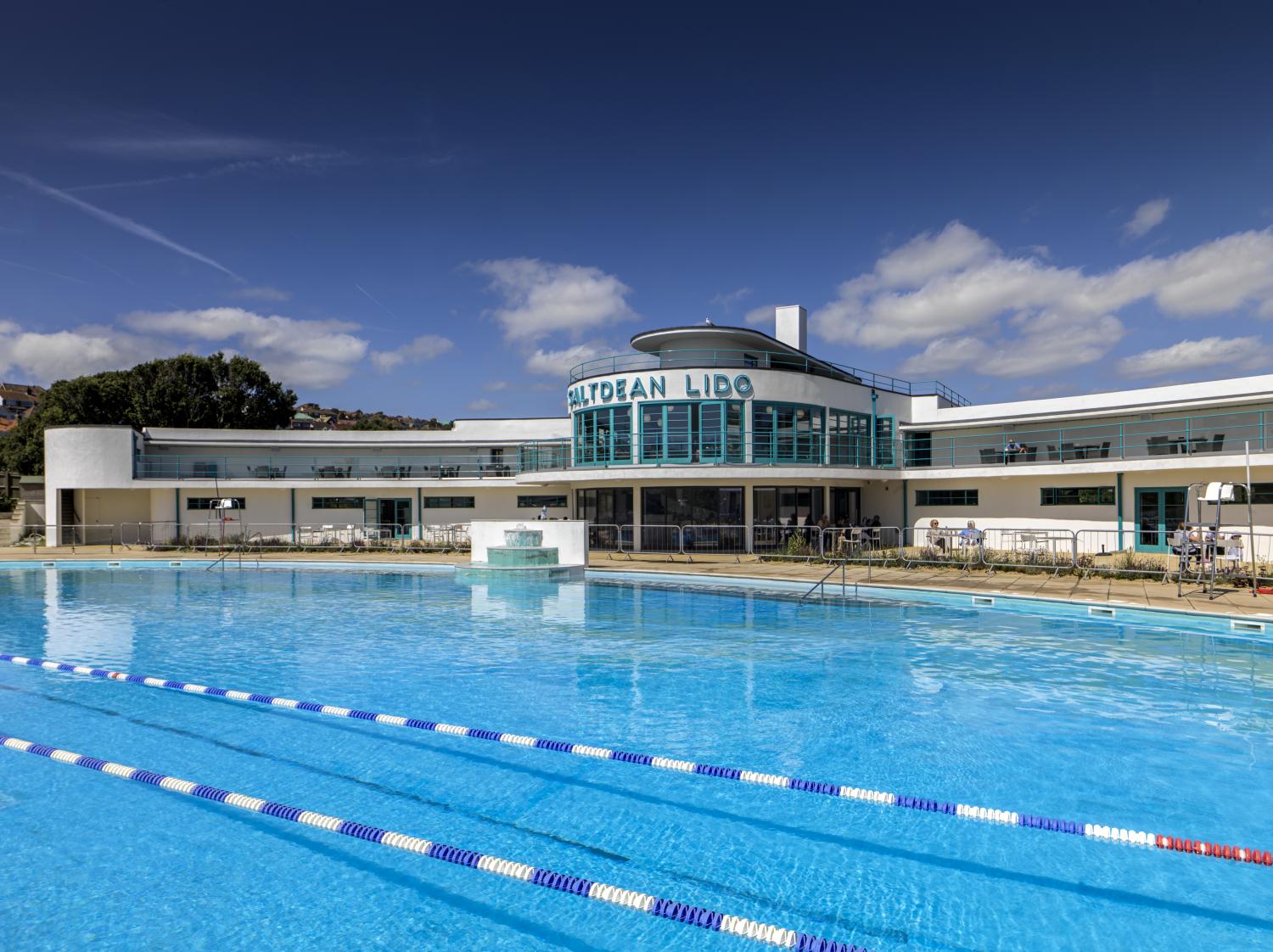 An exterior scenic photo of a newly renovated lido, with bright blue water in the foreground and cloudy blue skies above