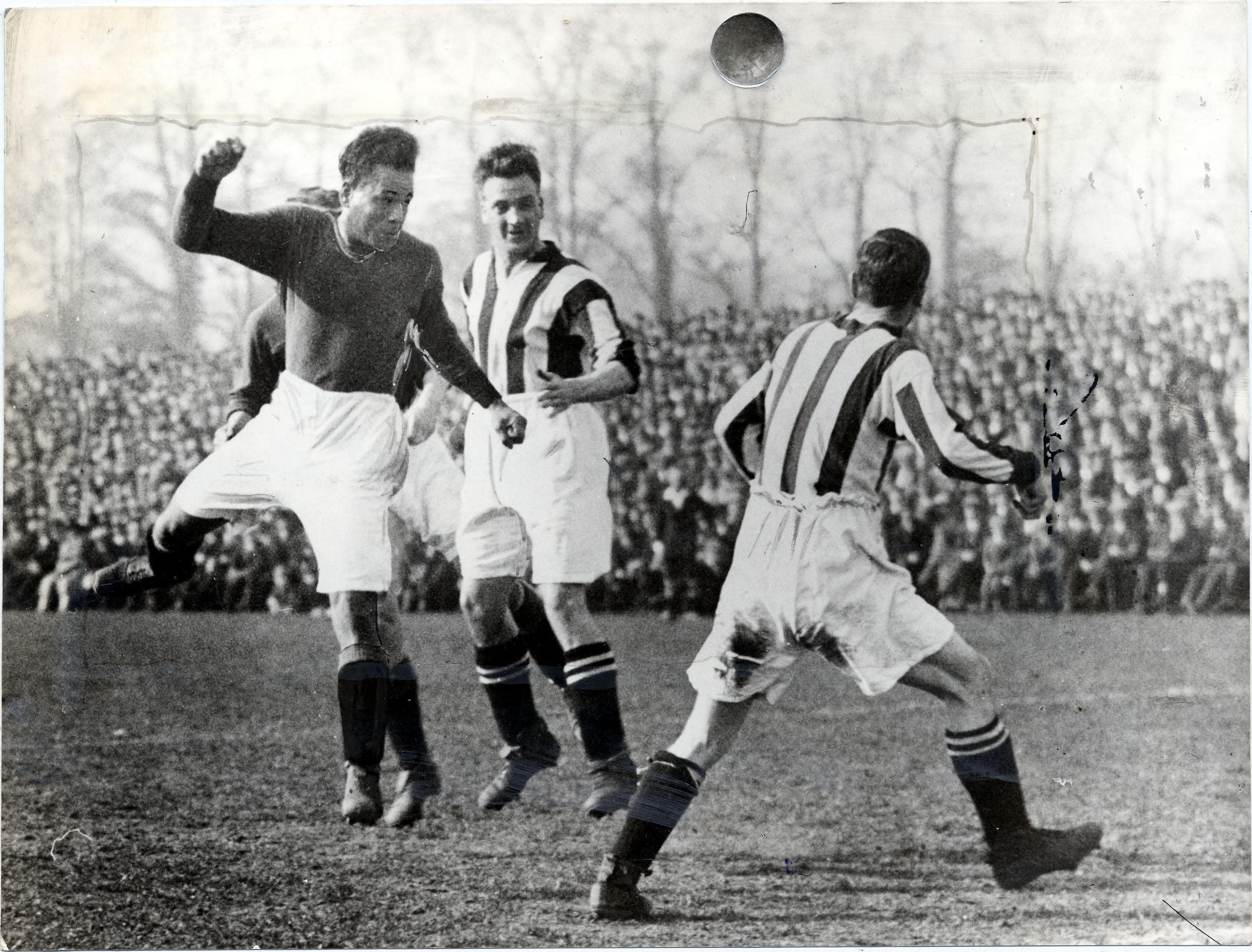A black and white photograph of a 1920s Black footballer leaping to head the ball, as two men in black and white striped shirts look on. A crowd is visible in the background.