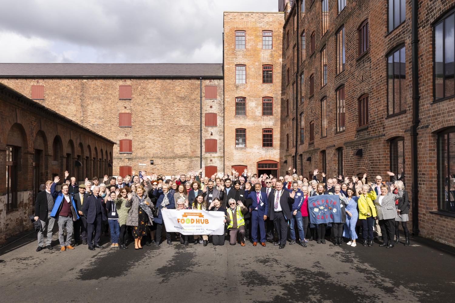 Group photograph of volunteers standing in a building forecourt smiling and waving for the camera. Some are holding banners.