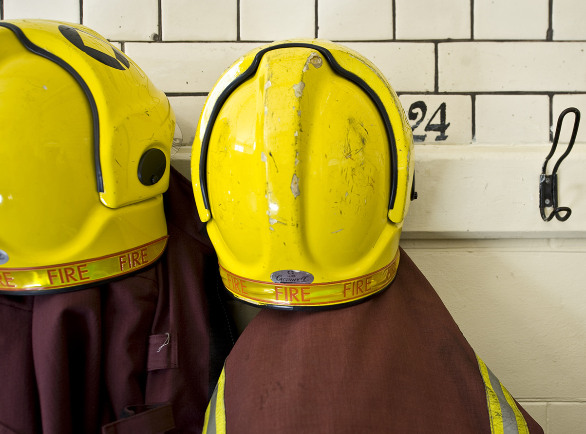 2 yellow helmets and maroon protective clothing hang on coat hooks in front of a white wall.